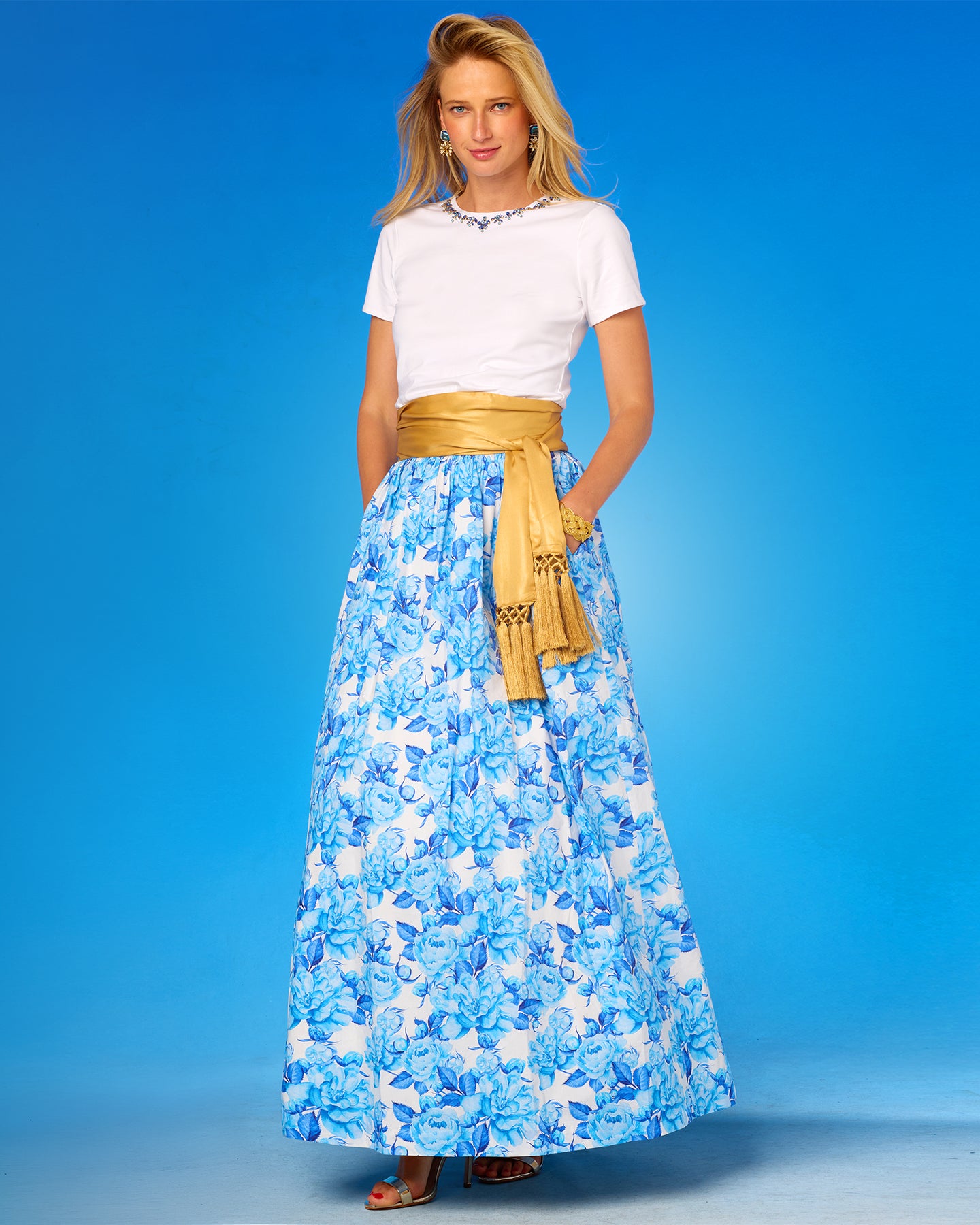 Woman wearing a white top and blue floral skirt with a gold belt against a blue background