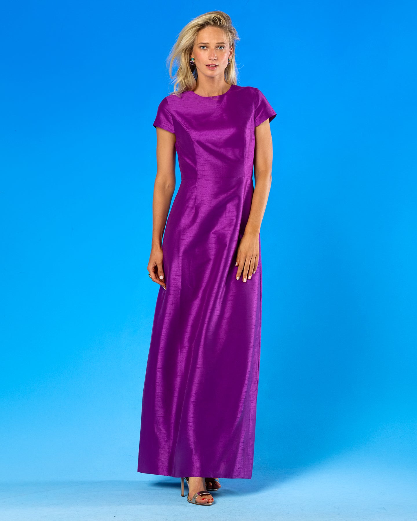 A woman modeling a floor-length, short-sleeve purple dress against a bright blue studio background.