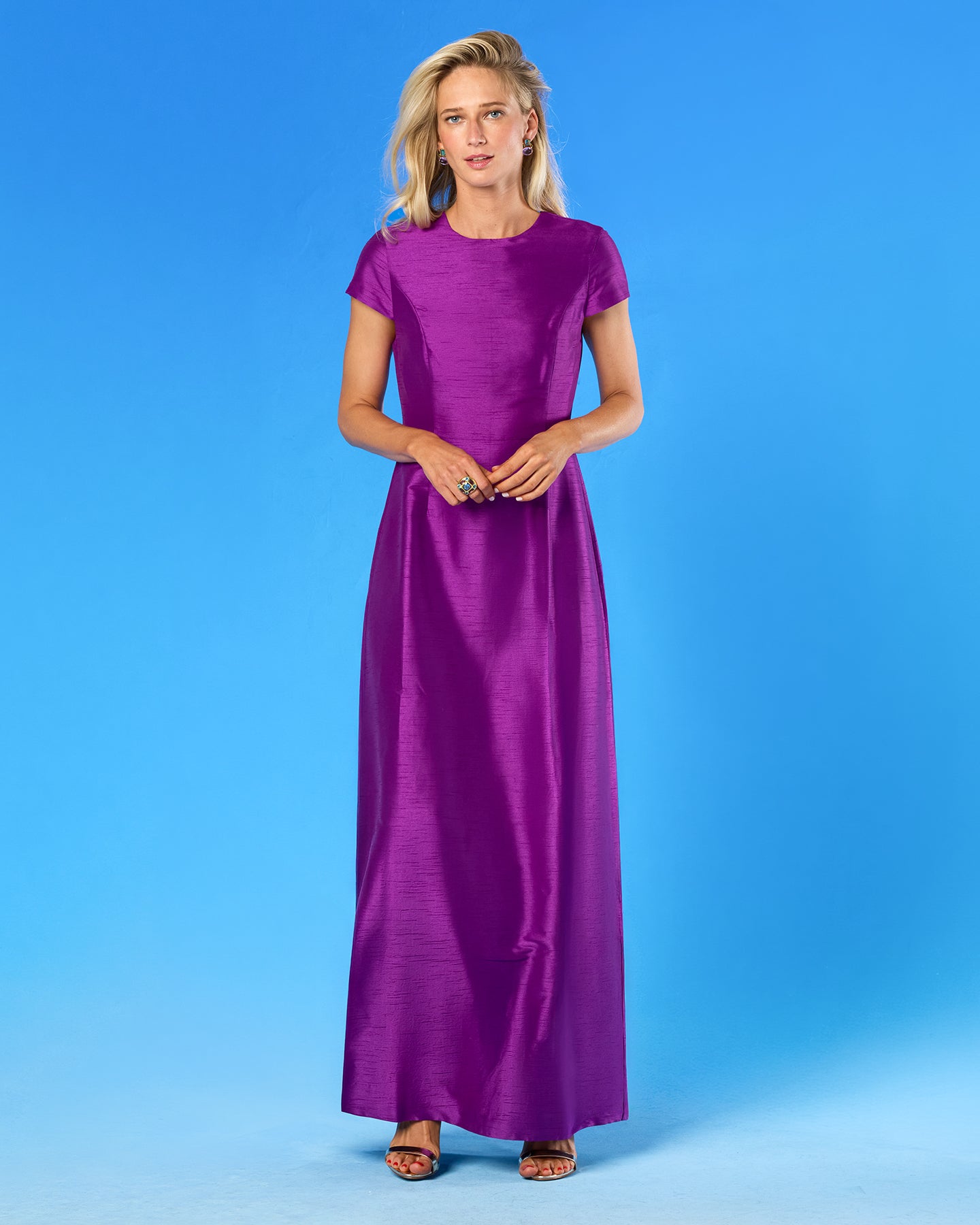 A woman modeling a long-sleeved, floor-length purple dress against a solid blue studio background.