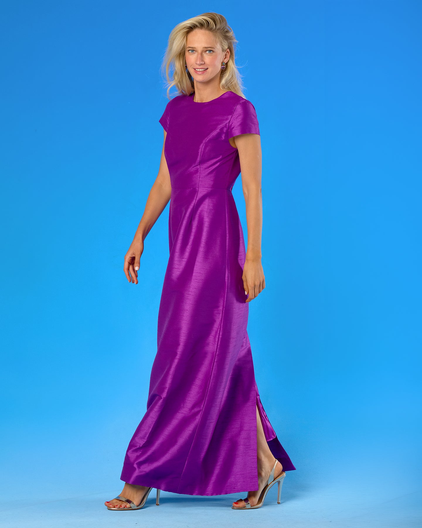 A woman modeling a floor-length magenta purple satin gown with short cap sleeves and a high side slit, standing against a solid blue studio background.
