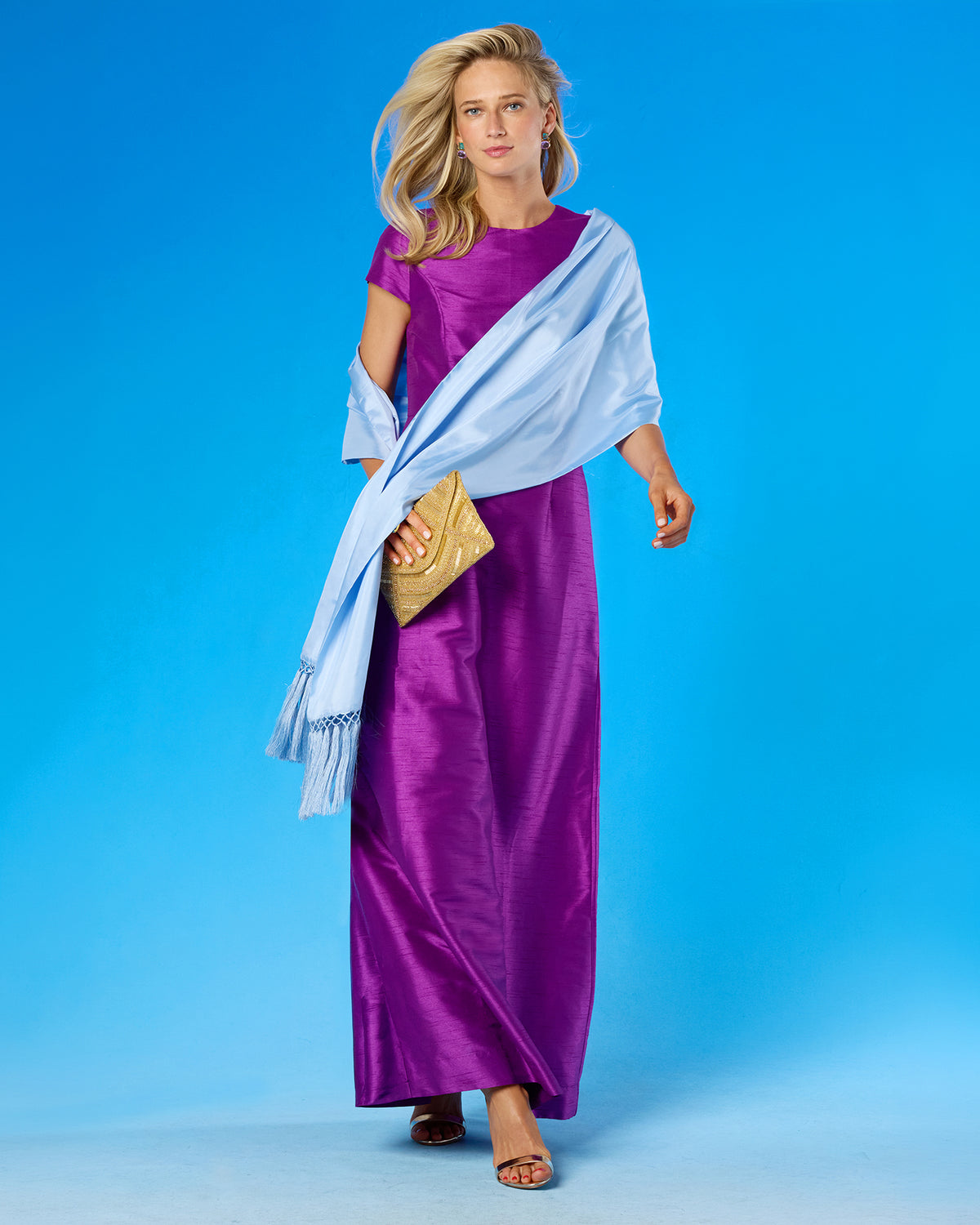 A woman modeling a long purple satin dress with a light blue draped shawl and a gold clutch, set against a solid blue studio background.