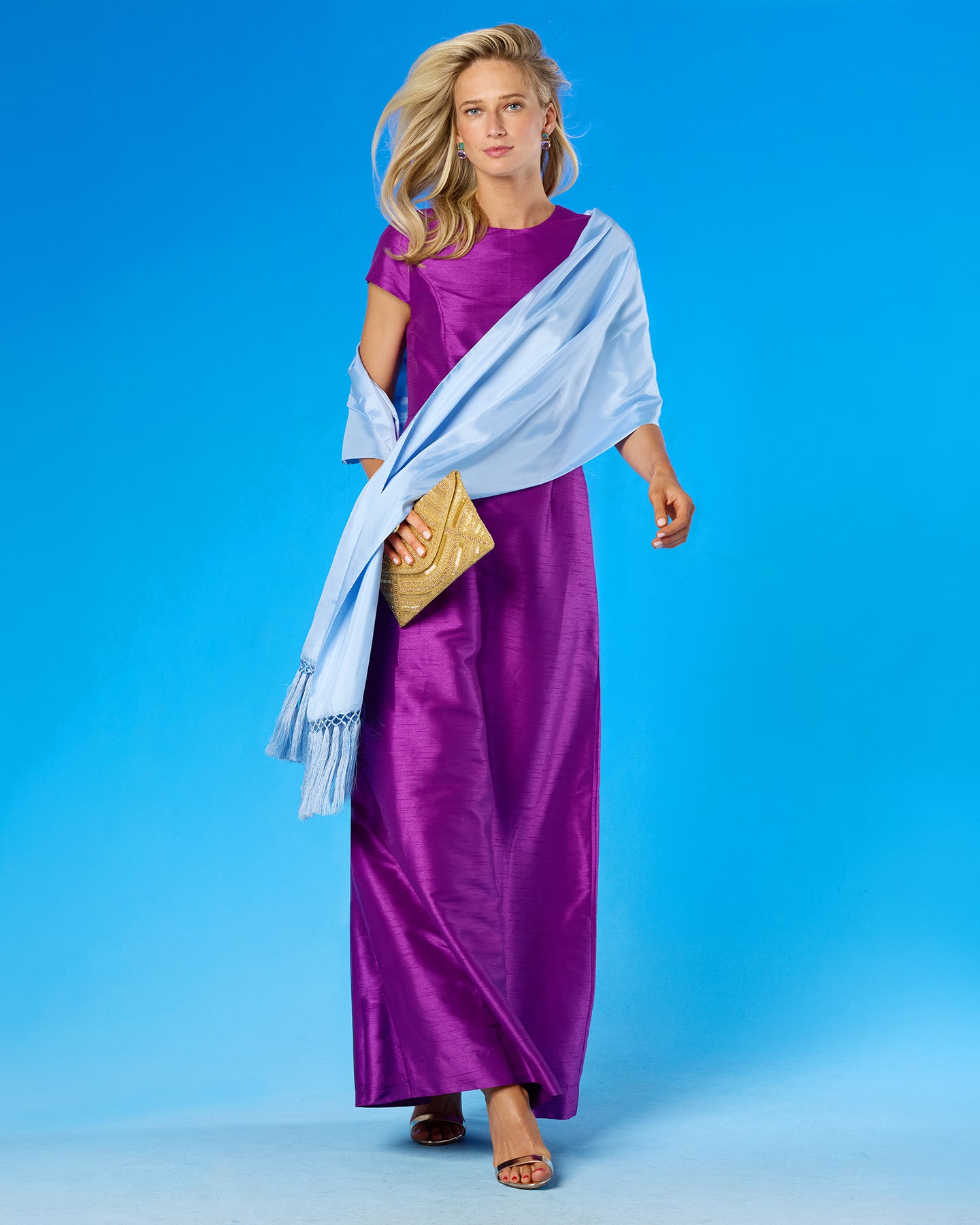A woman modeling a long purple satin dress with a light blue draped shawl and a gold clutch, set against a solid blue studio background.
