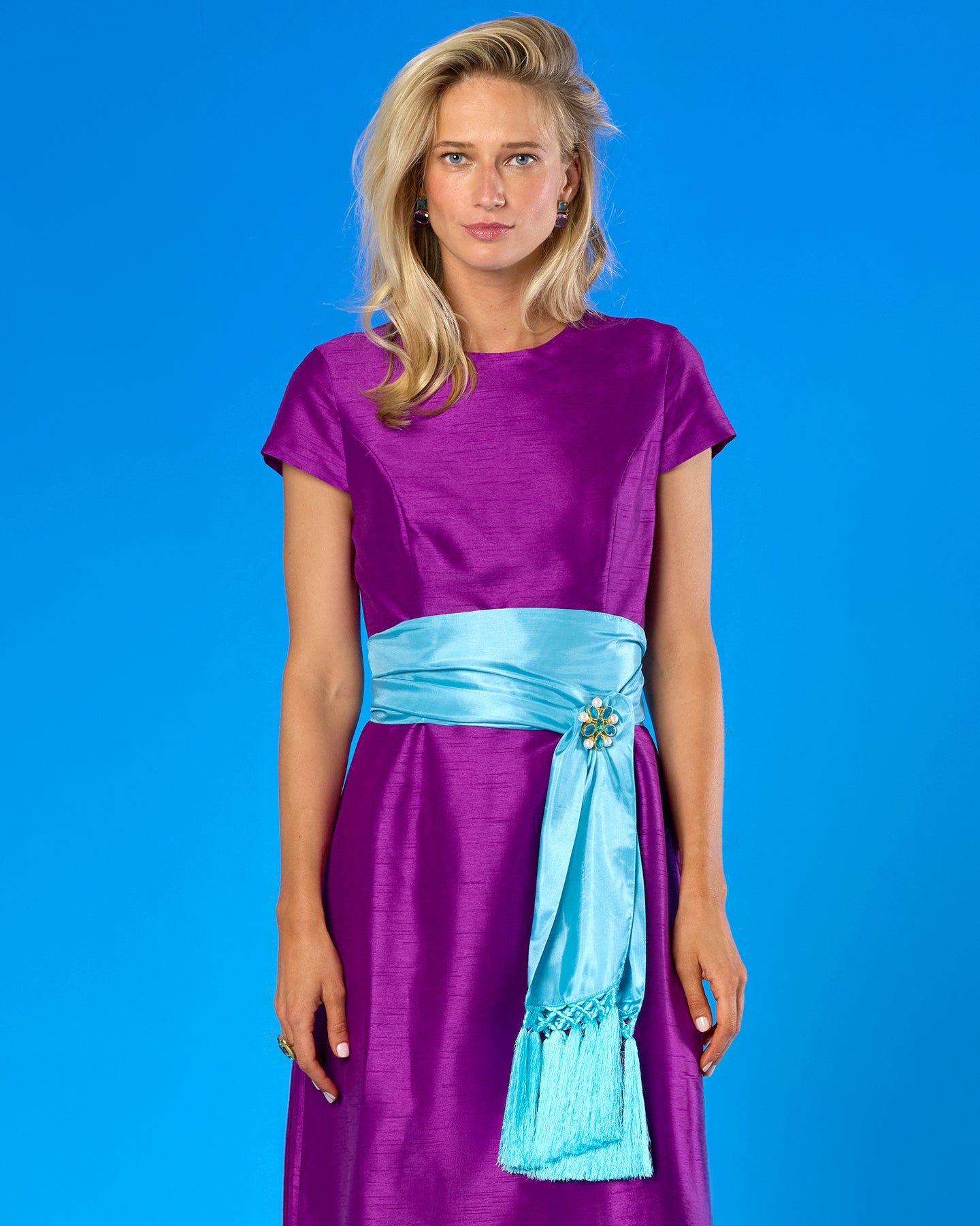 A woman modeling a purple short-sleeve dress with a light blue sash at the waist, set against a solid blue studio background.