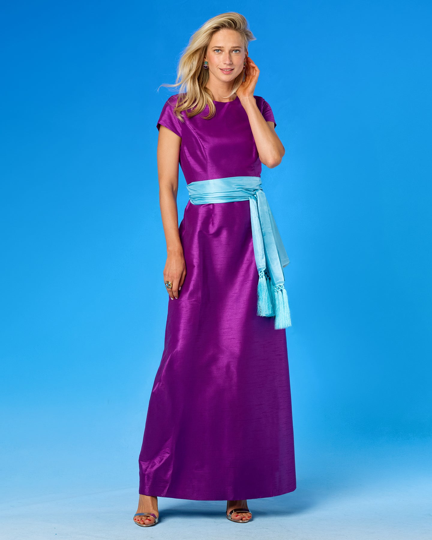 A woman modeling a floor-length purple satin dress with short cap sleeves and a light blue sash tied at the waist, posed against a solid blue studio background.