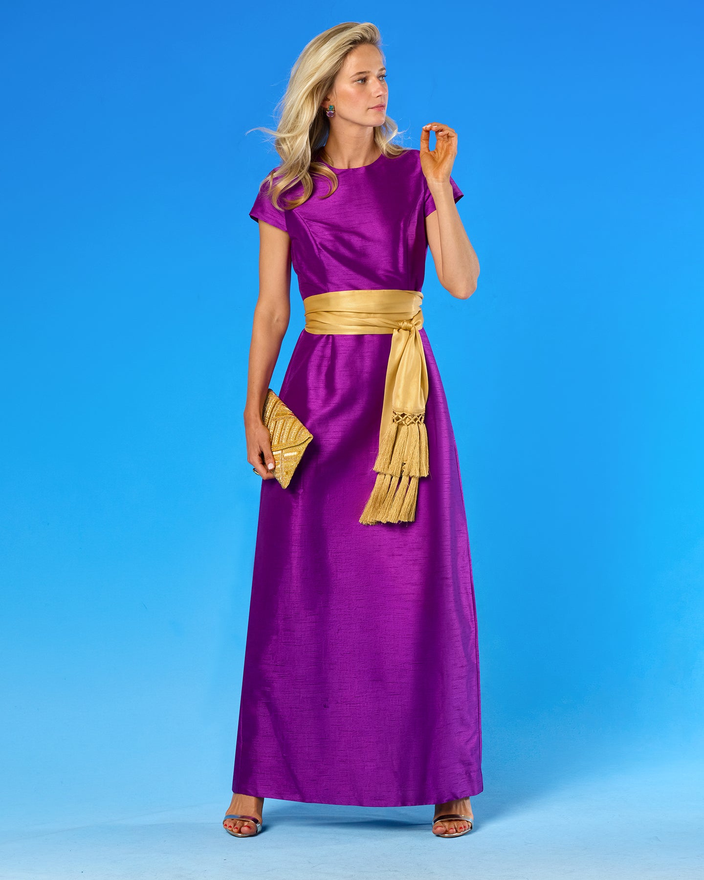 A woman modeling a floor-length purple dress with a wide gold sash at the waist, standing against a bright blue studio background.