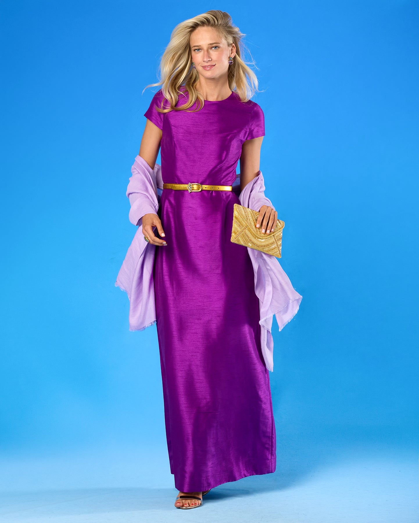 A woman modeling a floor-length purple gown with short sleeves, a gold belt, and draped lavender sleeves, standing against a bright blue studio background and holding a gold clutch.