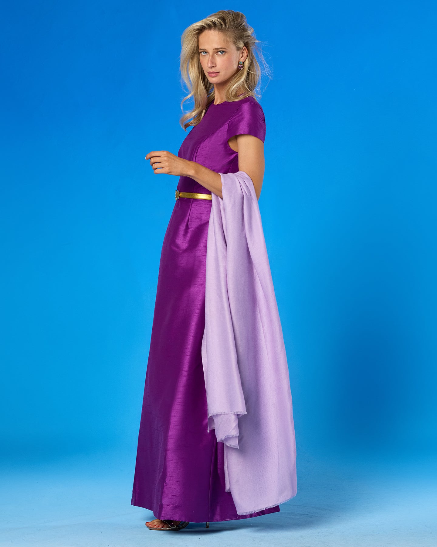 A woman modeling a floor-length purple satin gown with a gold waistband and a light lavender draped shawl, posed against a solid blue studio background.