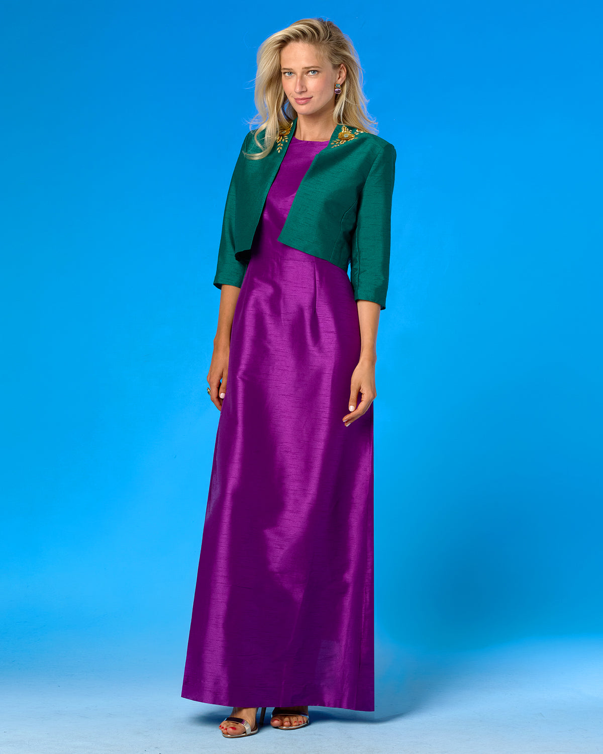 A woman modeling a formal outfit consisting of a long purple satin maxi dress and a dark green three-quarter sleeve bolero jacket with gold embroidery, set against a solid blue studio background.