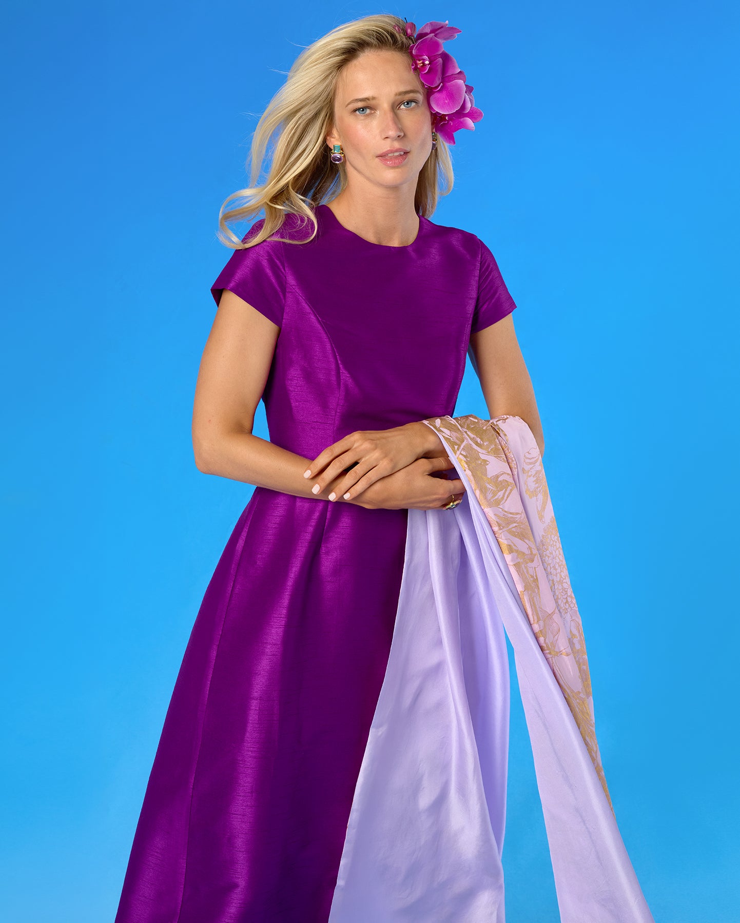 A woman modeling a short-sleeved magenta-purple satin dress with a matching lavender dupatta featuring gold embroidery, set against a solid blue studio background.
