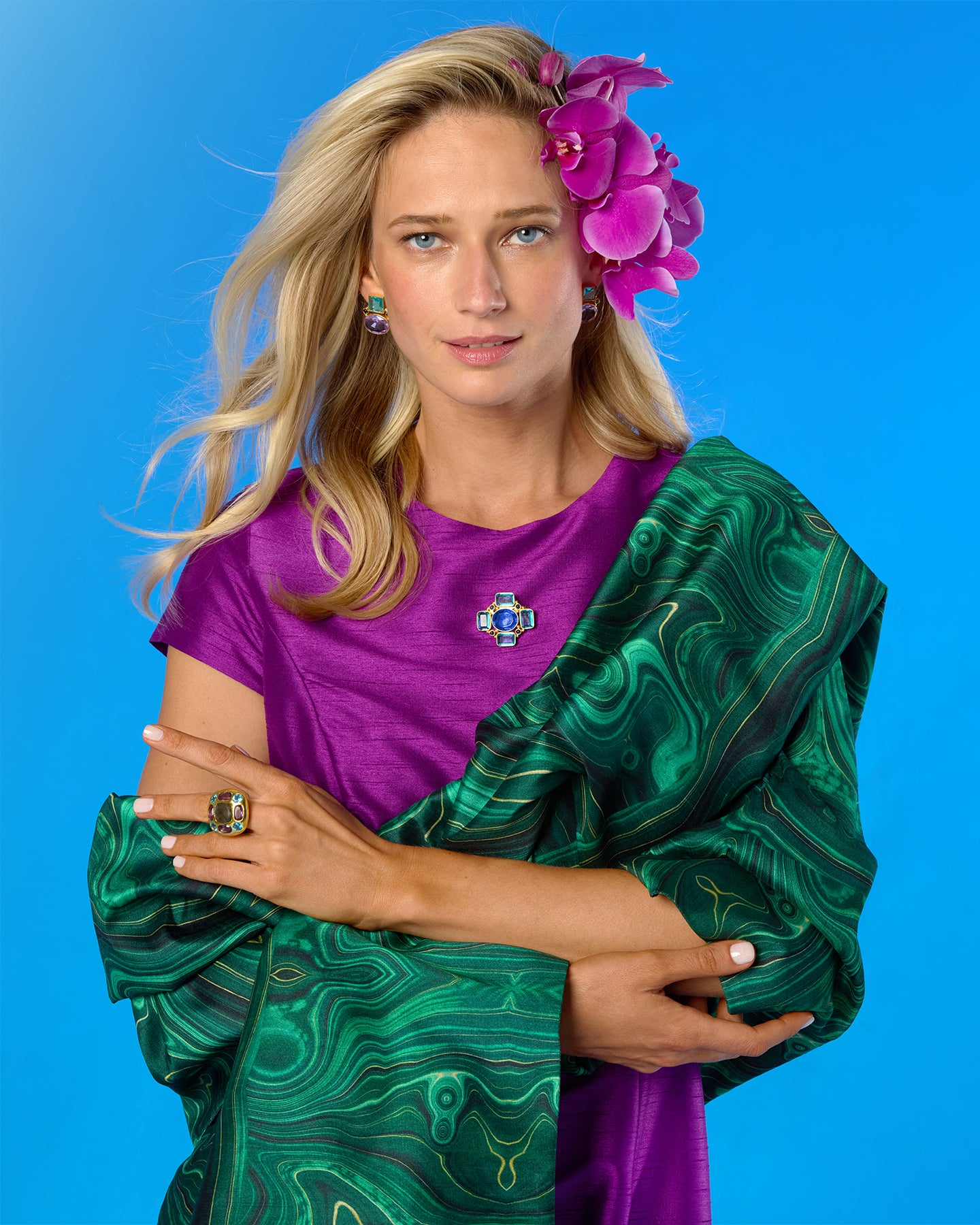 A woman with blonde hair and a purple flower accessory in her hair, wearing a purple top and a green marbled shawl, posing against a solid blue background.