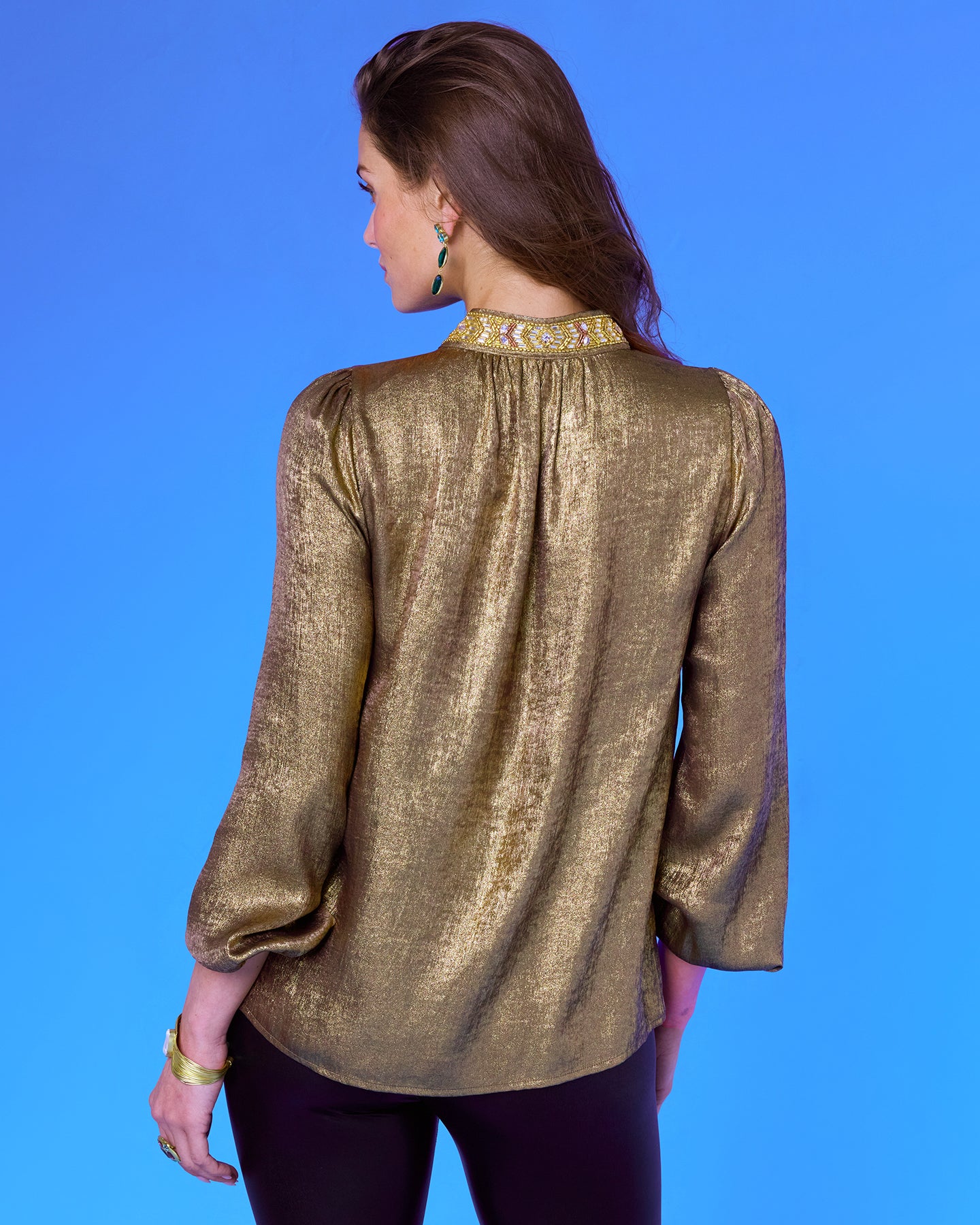 A woman viewed from the back wearing a metallic gold long-sleeve blouse with a patterned gold and white embroidered collar, set against a solid blue background.