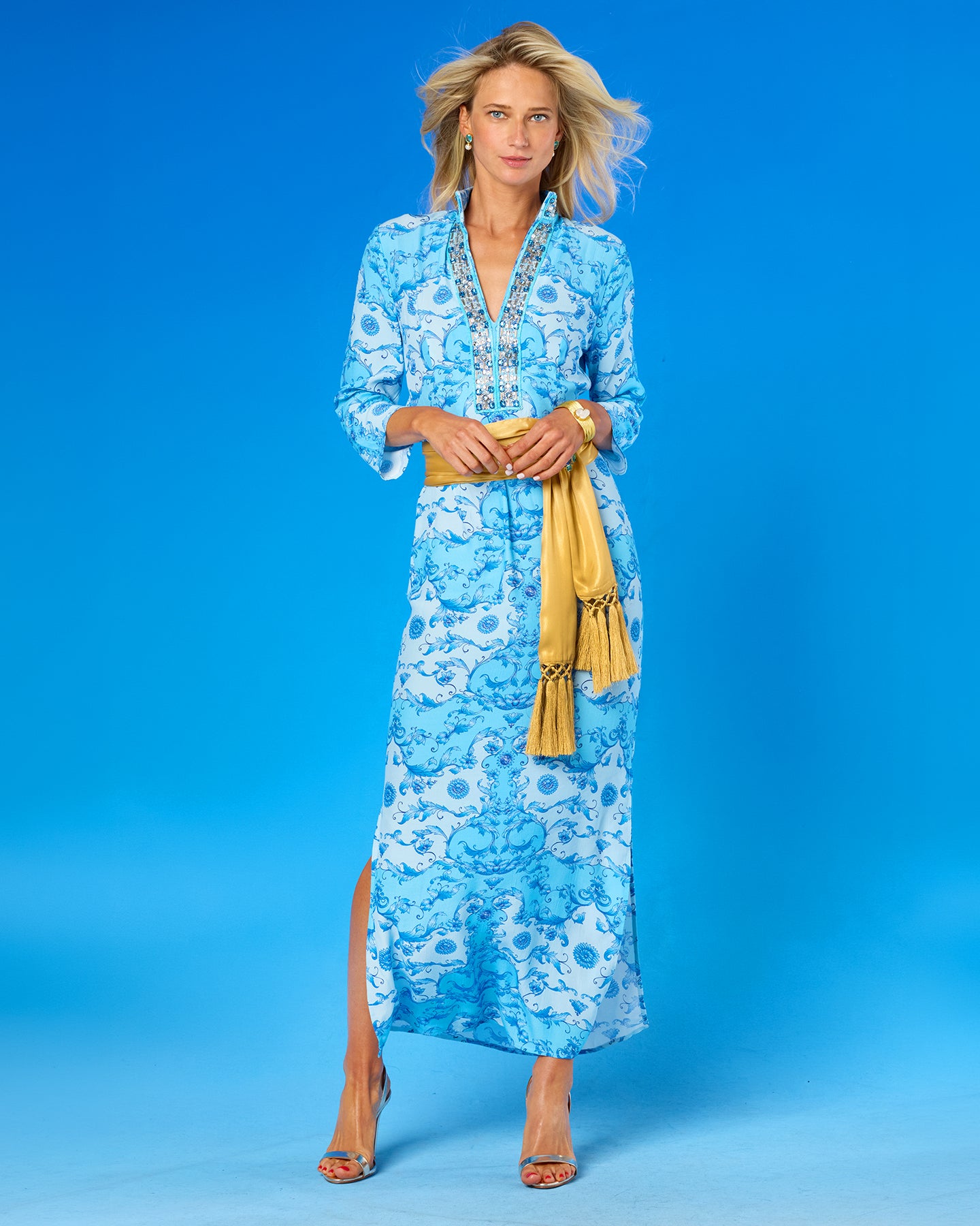 A woman modeling a long-sleeve blue and white patterned maxi dress with a yellow tassel belt, standing against a solid blue studio background.