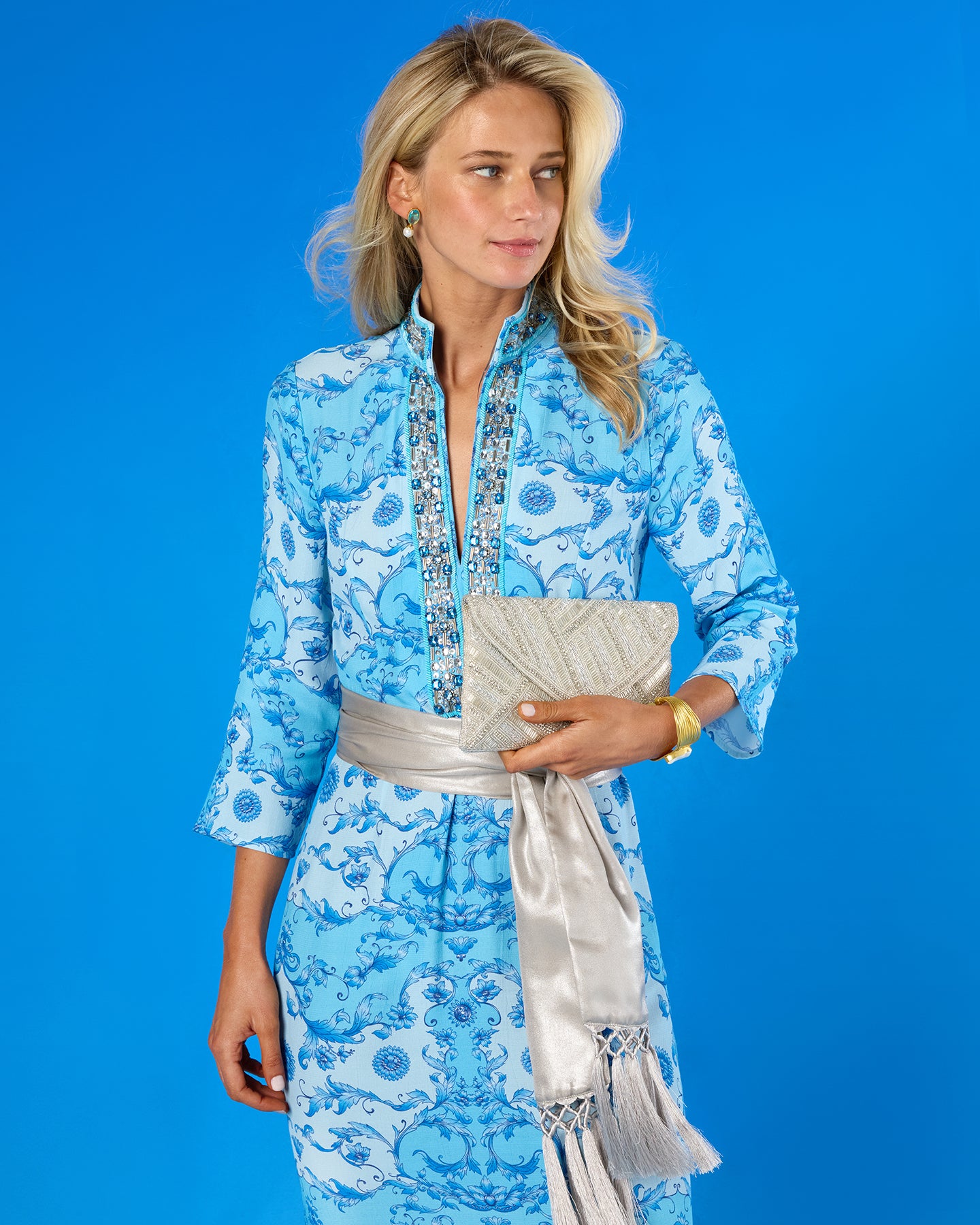 A woman modeling a light blue and white patterned dress with a silver belt and clutch, set against a solid blue studio background.