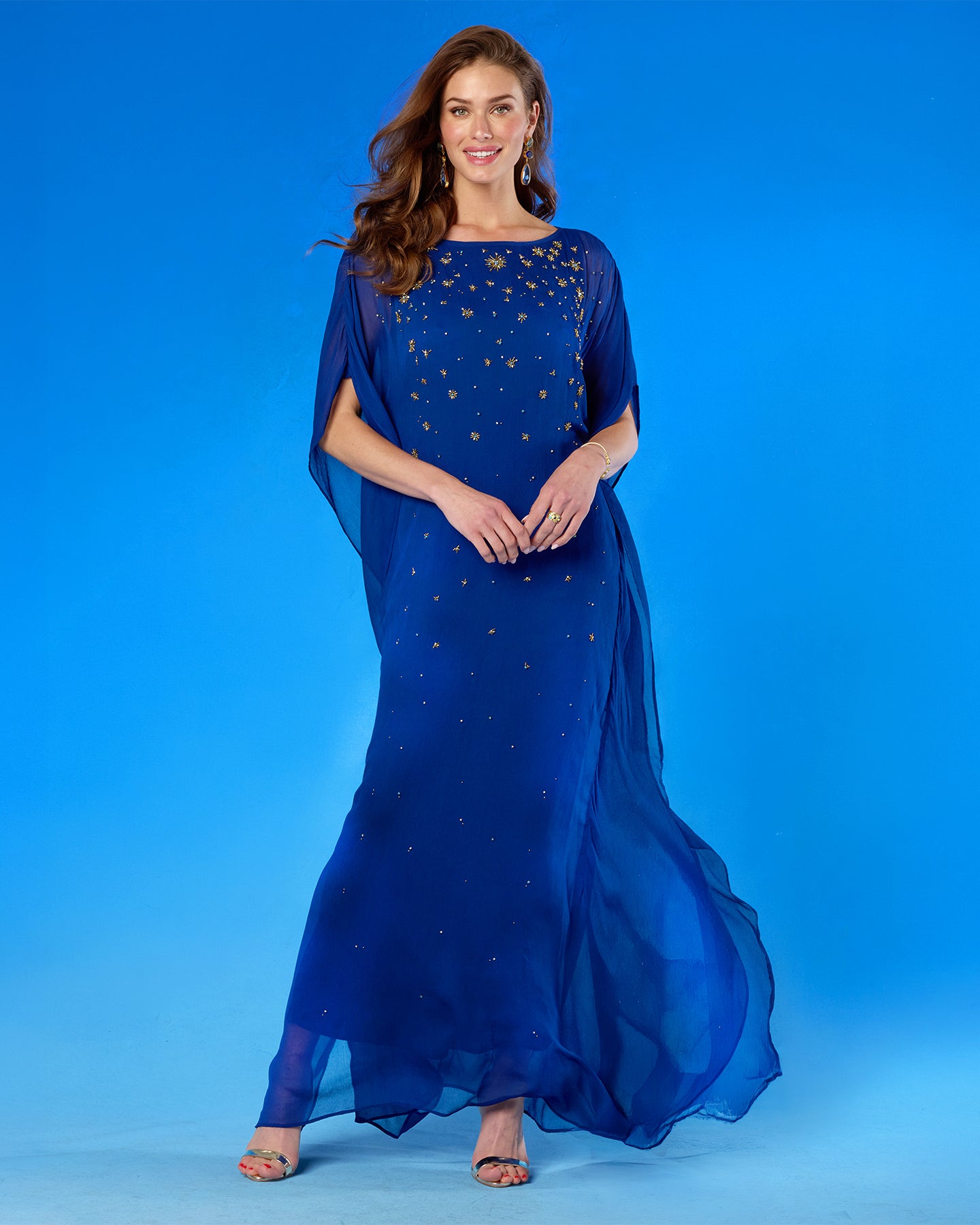 A woman modeling a floor-length royal blue gown with sheer draped cap sleeves and scattered gold star-like embellishments, posed against a solid blue studio backdrop.