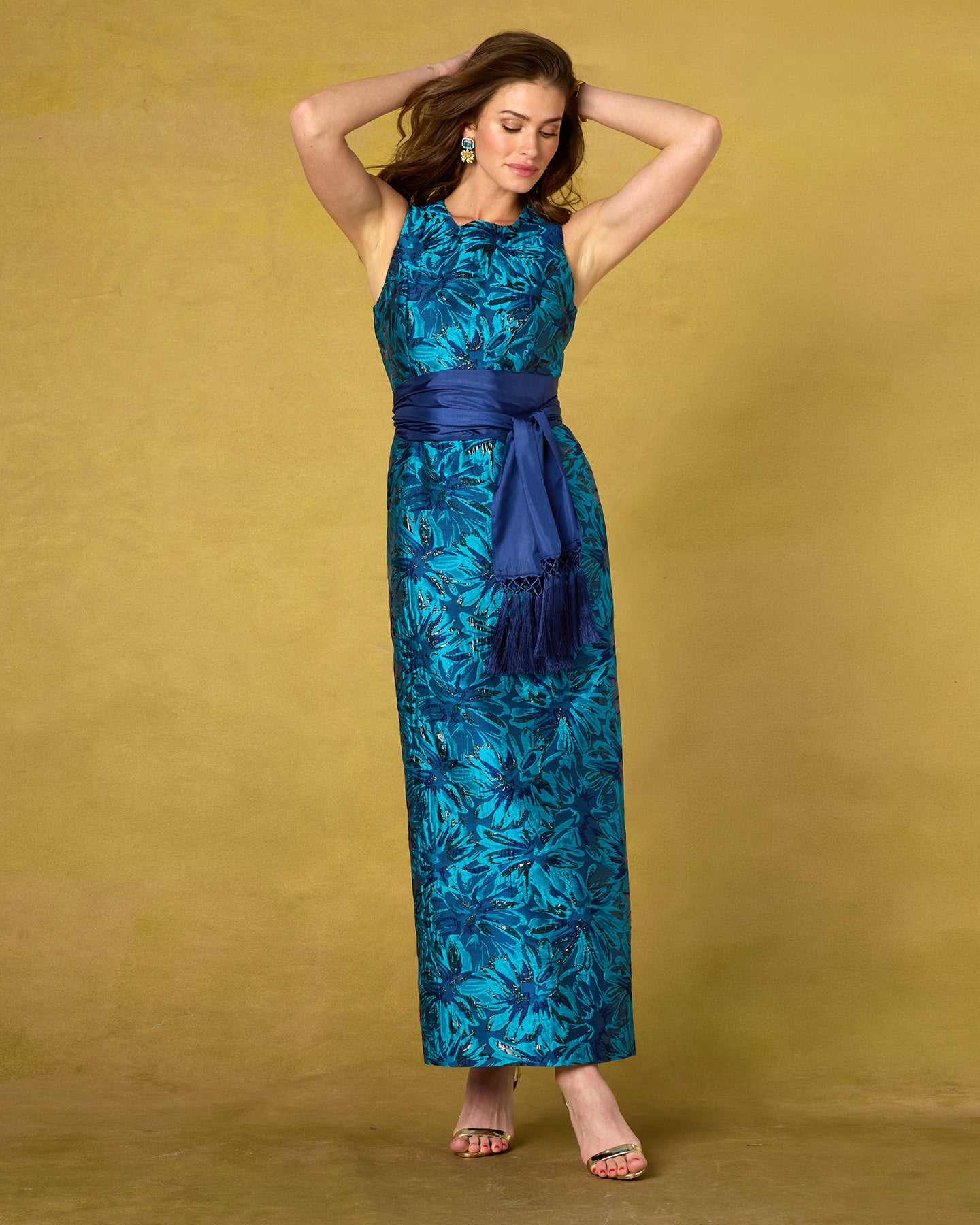A woman modeling a sleeveless maxi dress with a blue and teal floral pattern and a navy blue sash, standing against a textured mustard-yellow studio backdrop.