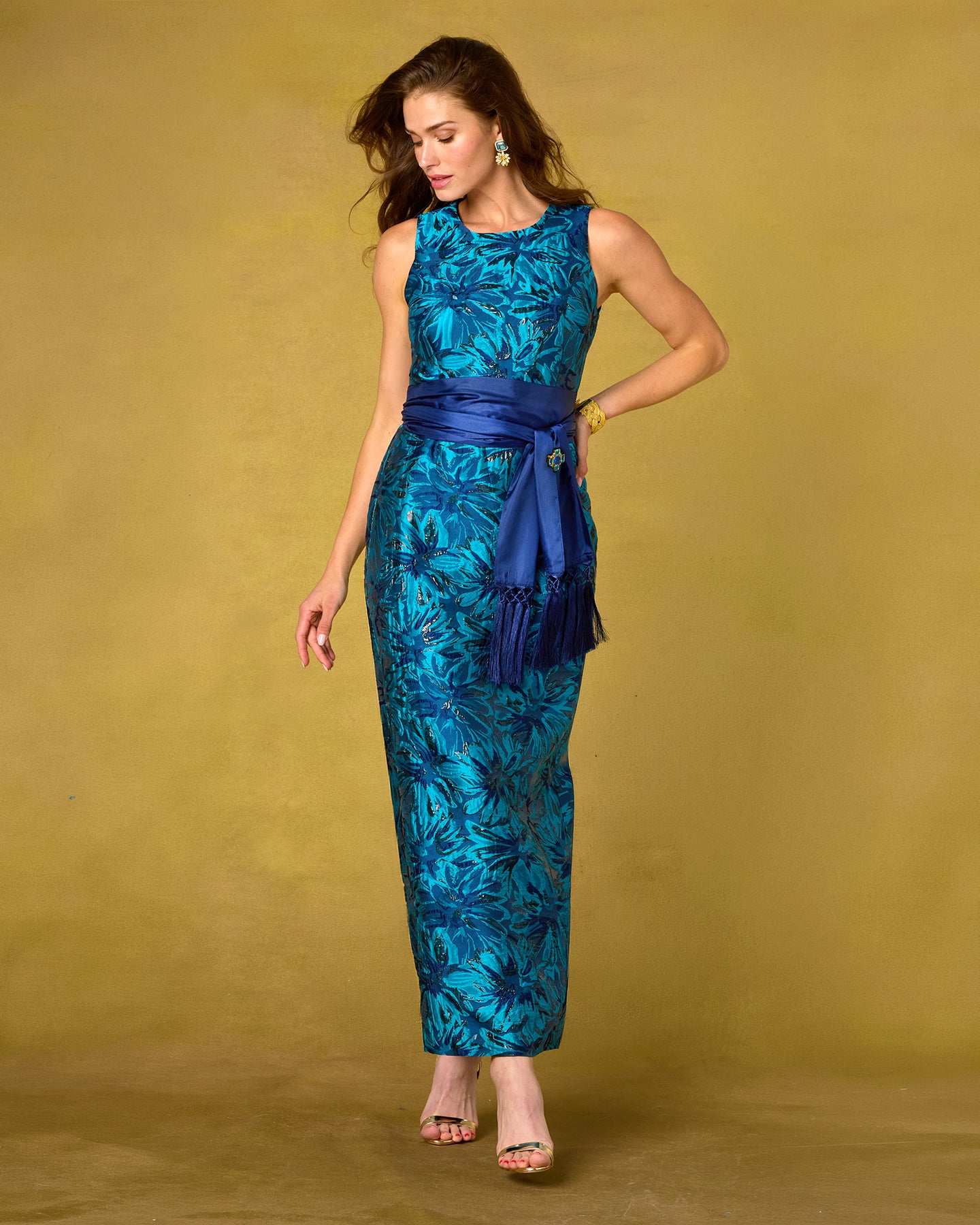 A woman models a sleeveless, floor-length teal and blue tropical floral print dress with a matching dark blue sash, posed against a solid mustard yellow studio backdrop.
