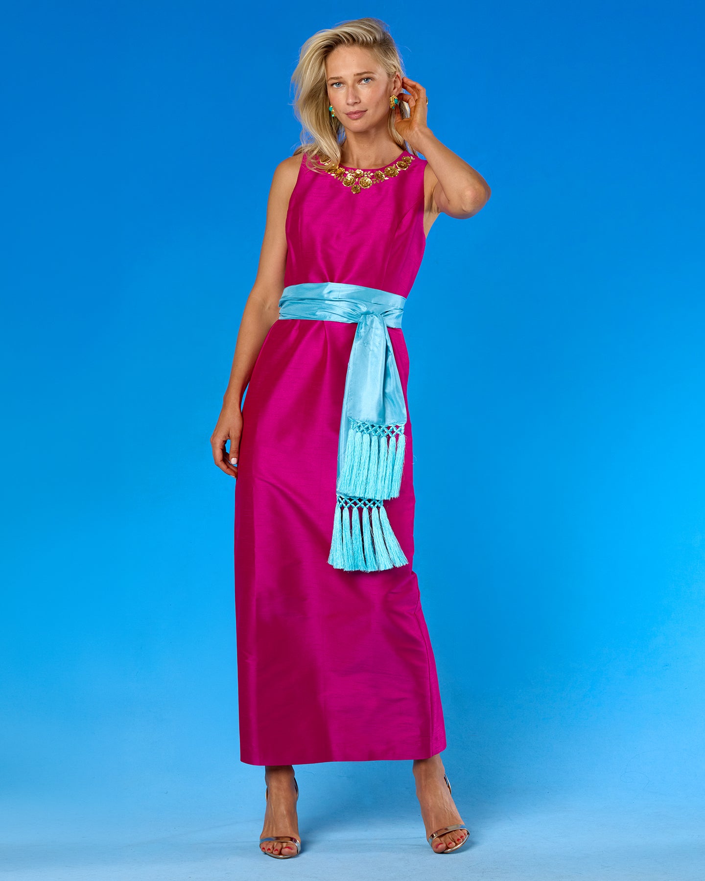 A woman modeling a sleeveless magenta maxi dress with a light blue sash and tassel detail, set against a solid blue studio background.