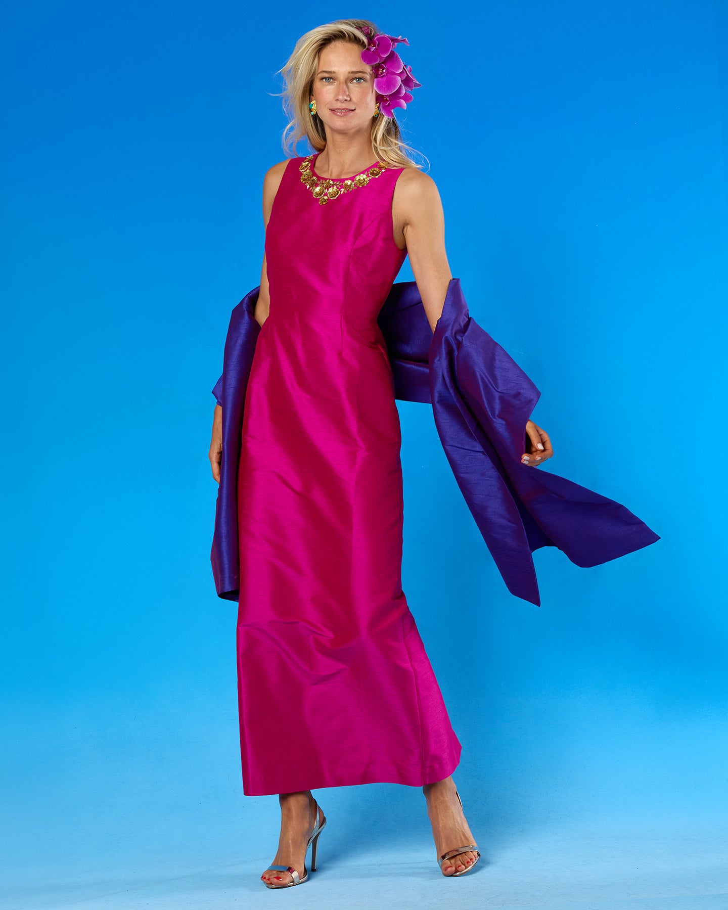 A woman models a sleeveless magenta maxi dress with a draped purple shawl over her shoulders, set against a solid blue studio background.