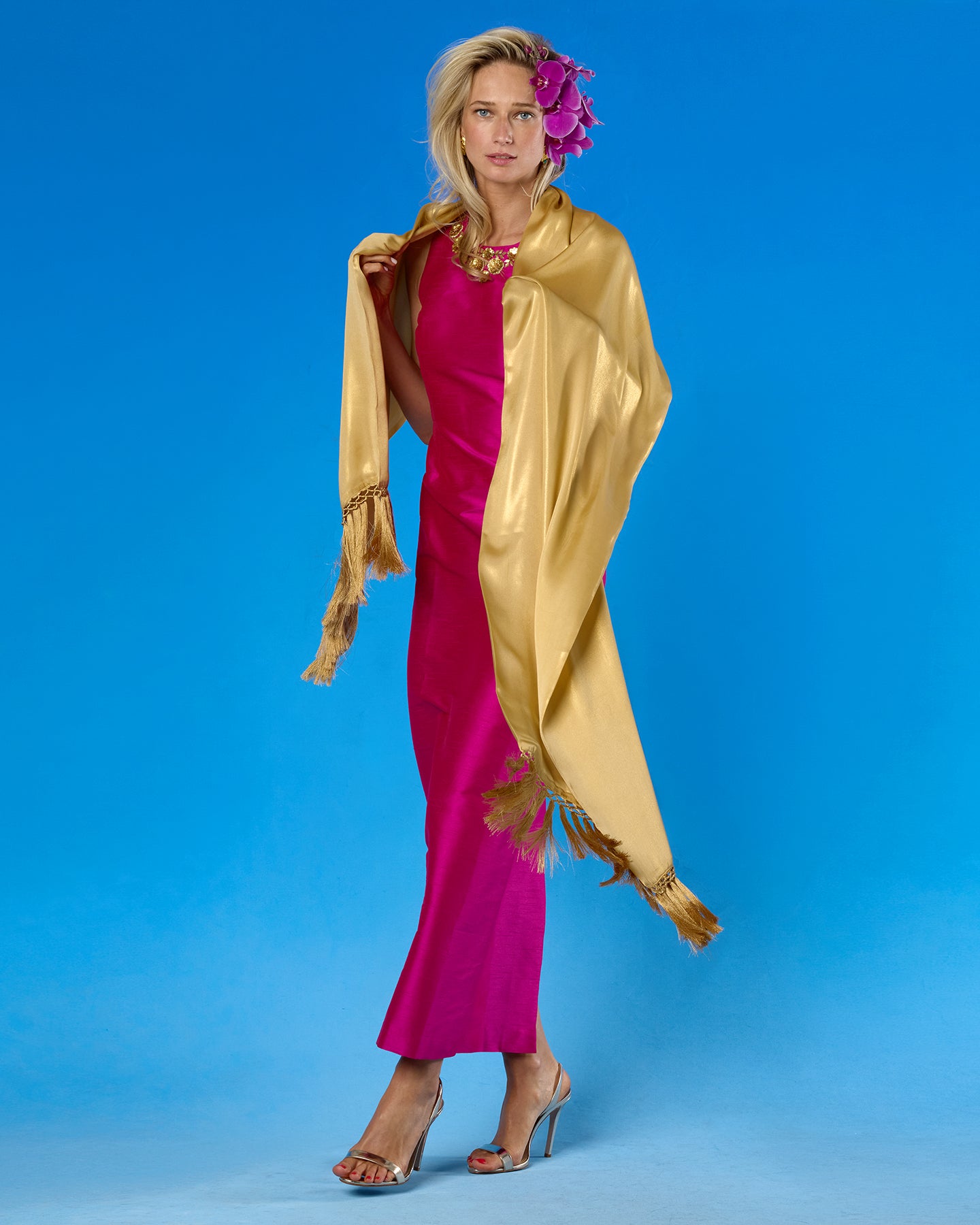 A woman models a two-tone dress with one side in magenta pink and the other in a shiny gold satin wrap, styled with a gold necklace, gold heels, and a purple flower in her hair against a solid blue studio background.