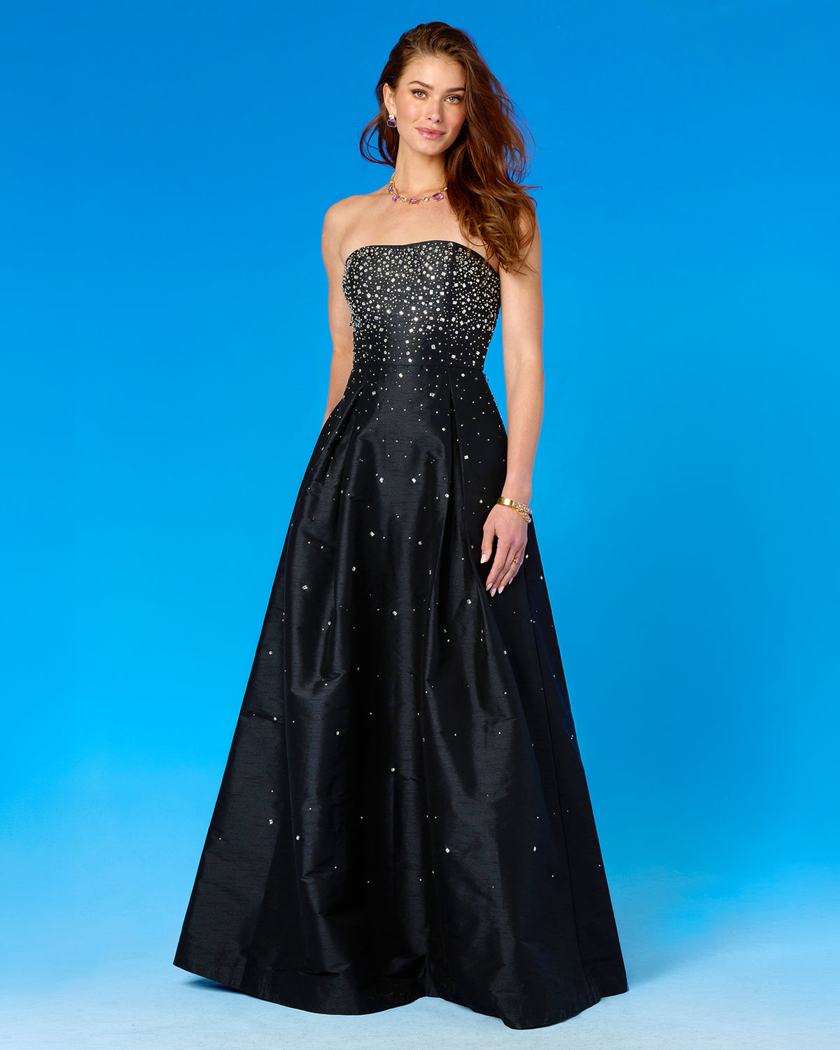 A woman modeling a strapless black A-line formal gown with a heavily embellished beaded bodice and scattered rhinestone-like accents on the skirt, posed against a bright blue studio background.