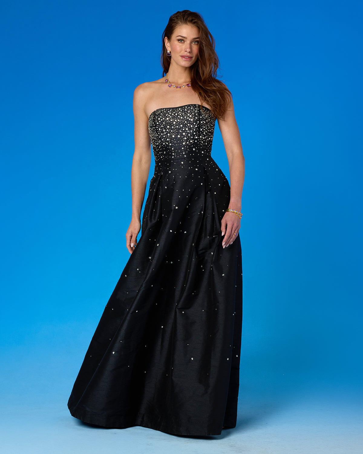 A woman models a strapless black taffeta gown embellished with sparkling silver rhinestones, set against a solid blue studio background.