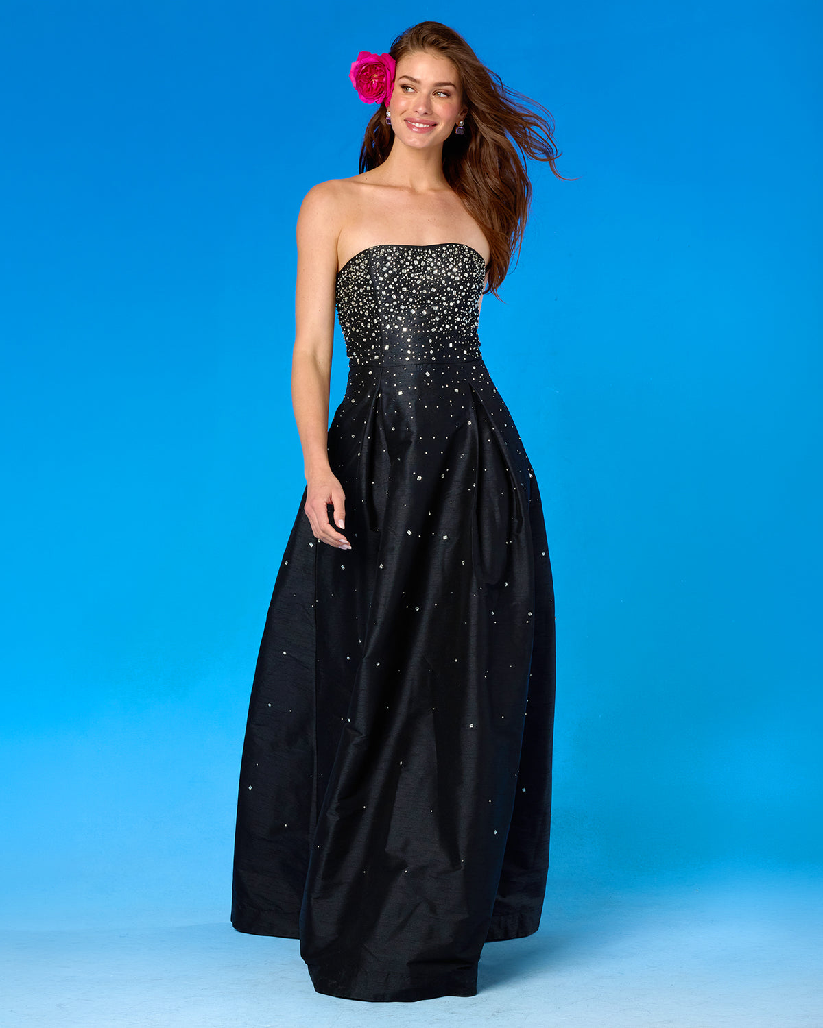 A woman modeling a strapless black taffeta formal gown with a heavily embellished beaded bodice and scattered rhinestone details on the skirt, set against a solid blue studio background.