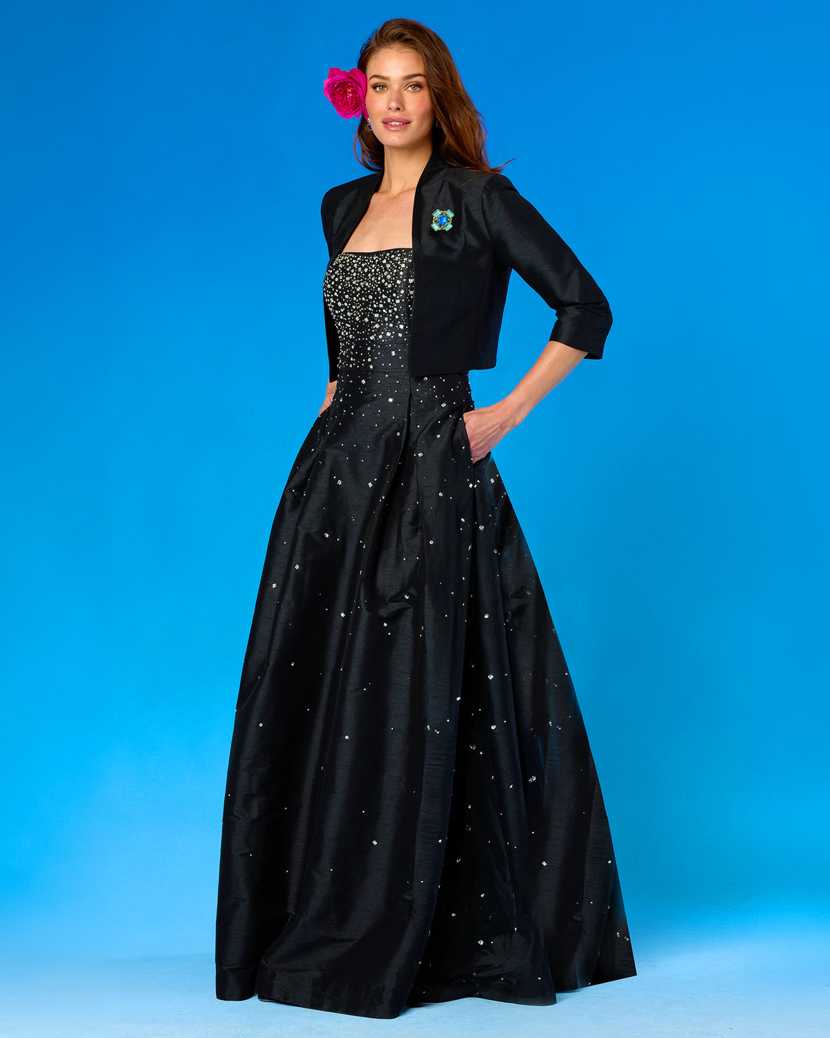 A woman models a formal black gown with a beaded bodice and scattered rhinestone embellishments, paired with a matching black jacket, against a bright blue studio background.
