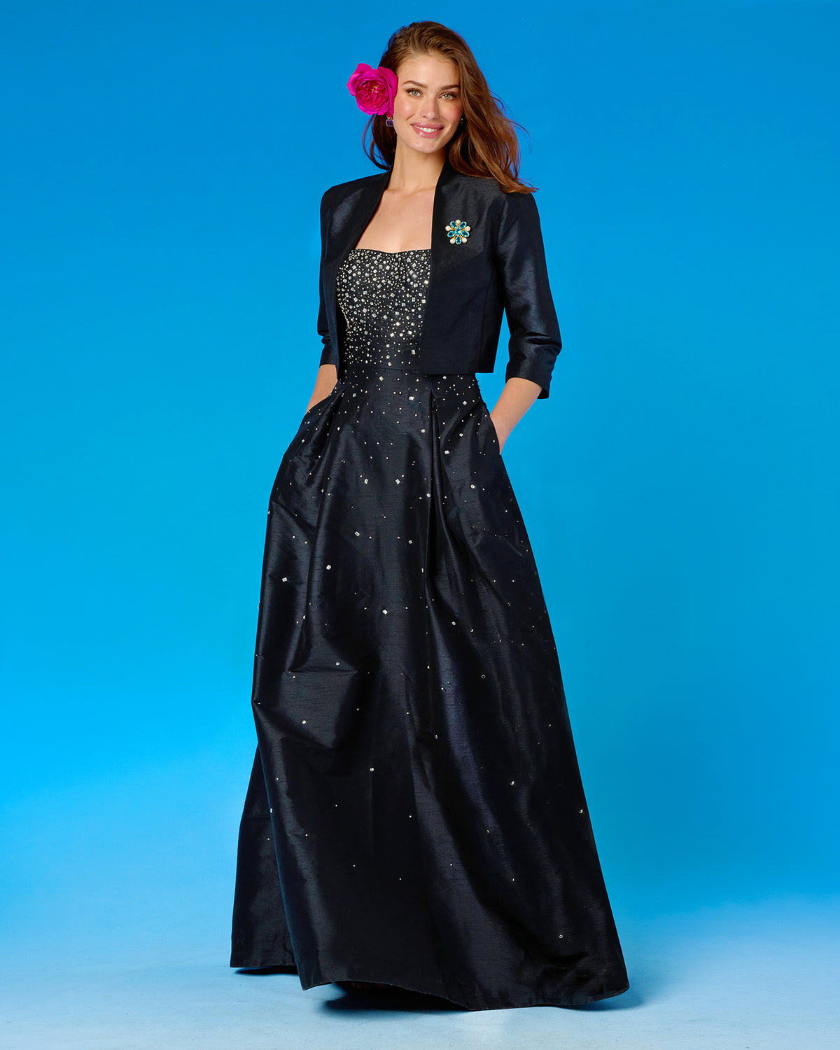 A woman modeling a formal black gown with a sparkling embellished bodice, a matching black jacket, and a bright pink flower accessory in her hair, set against a solid blue studio background.