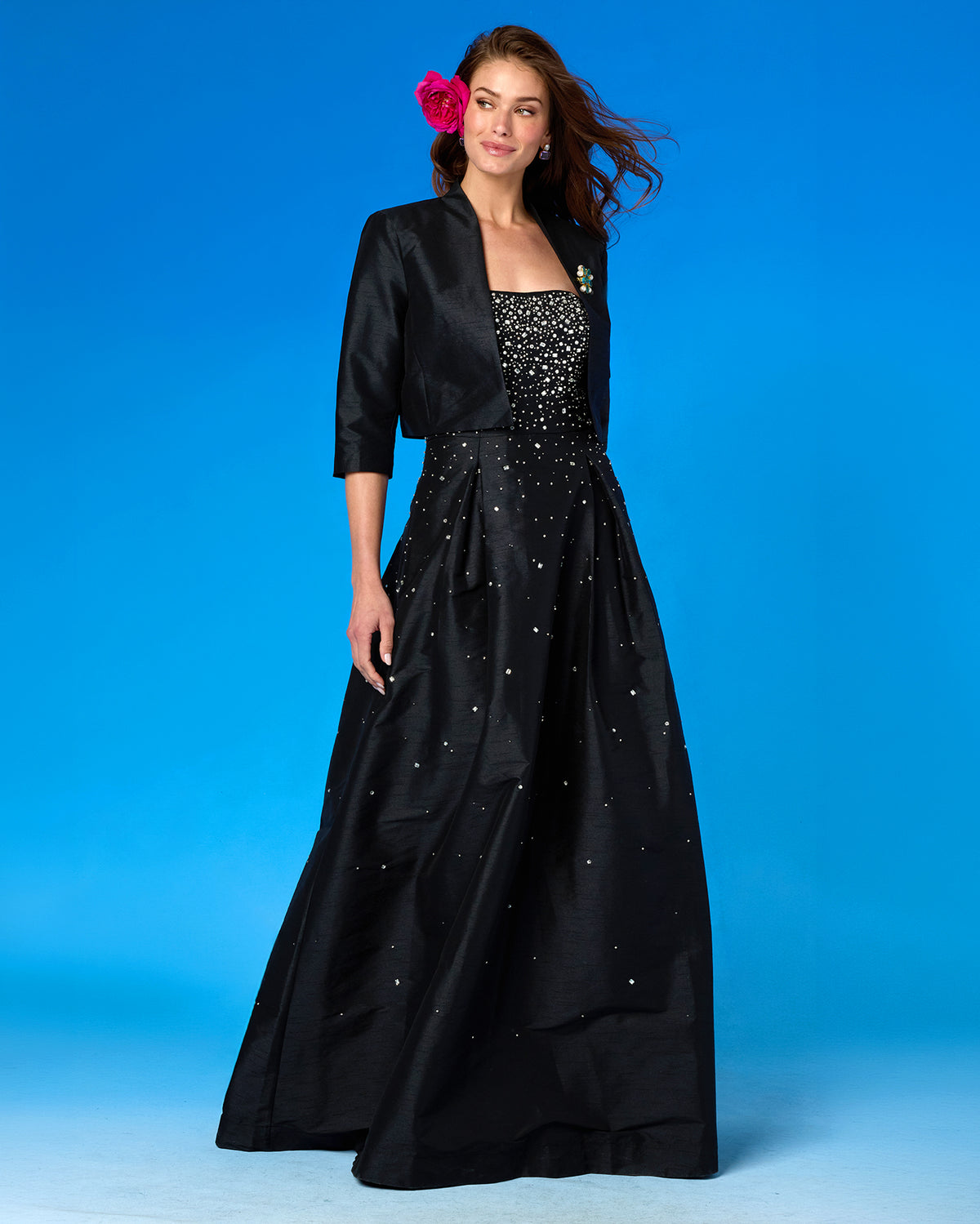 A woman modeling a formal black gown with a matching three-quarter sleeve bolero jacket. The dress features a beaded bodice and scattered rhinestone embellishments on the skirt, set against a solid blue studio background.