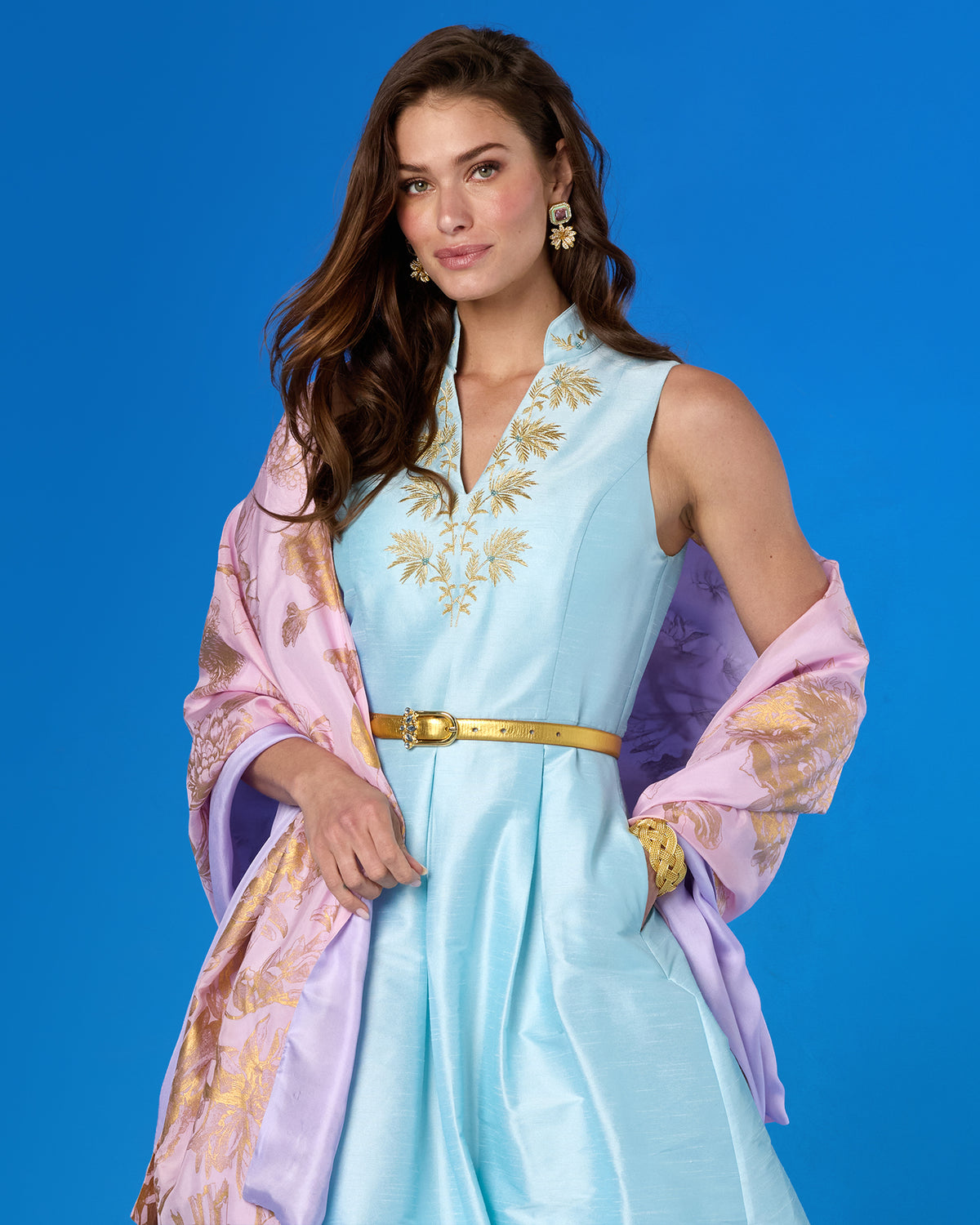 A woman modeling a sleeveless light blue dress with gold embroidery, a gold belt, and a lavender and gold patterned shawl, posed against a solid blue studio background.