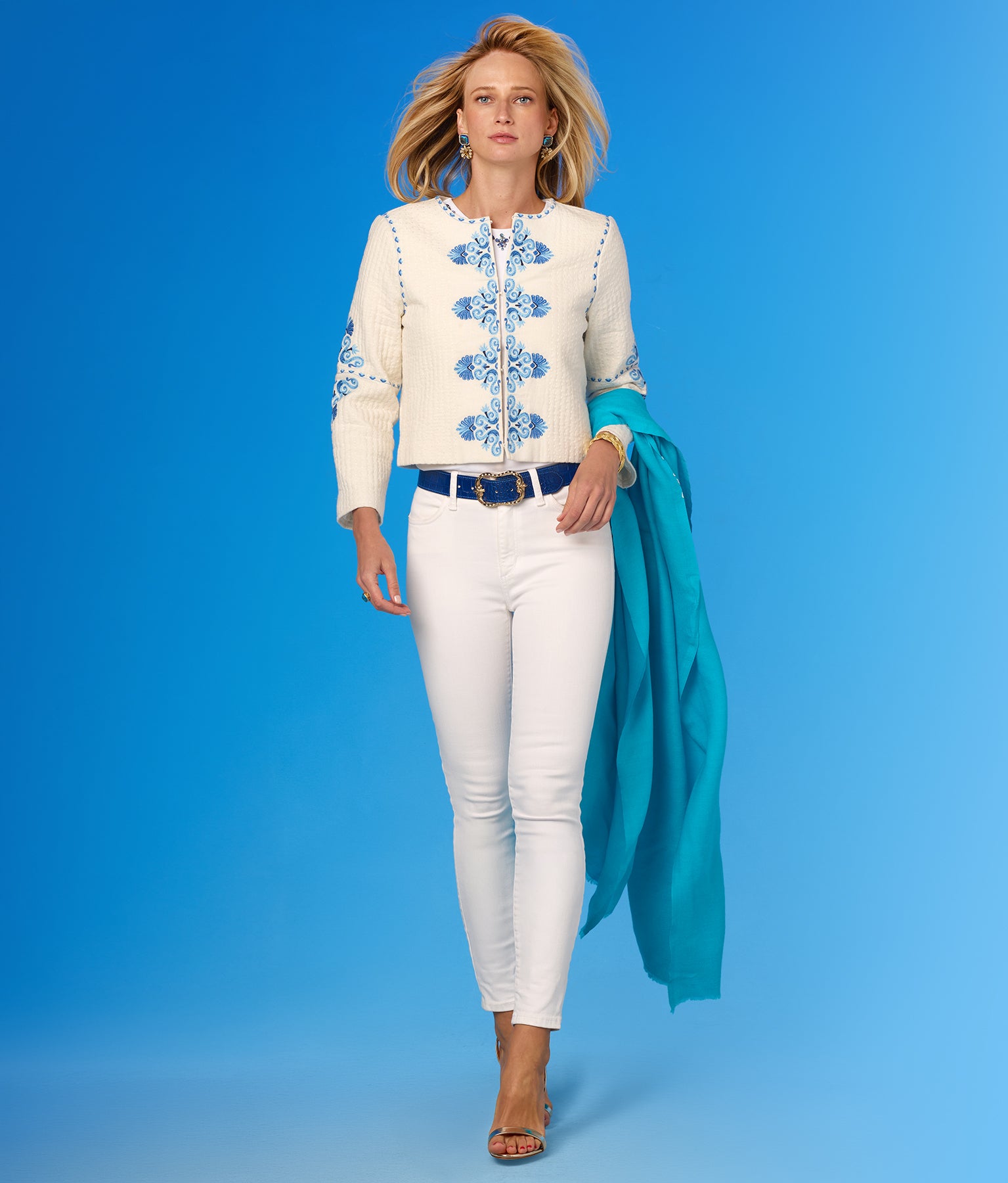 Woman wearing a white embroidered jacket with blue patterns, white pants, and a turquoise scarf on a blue background