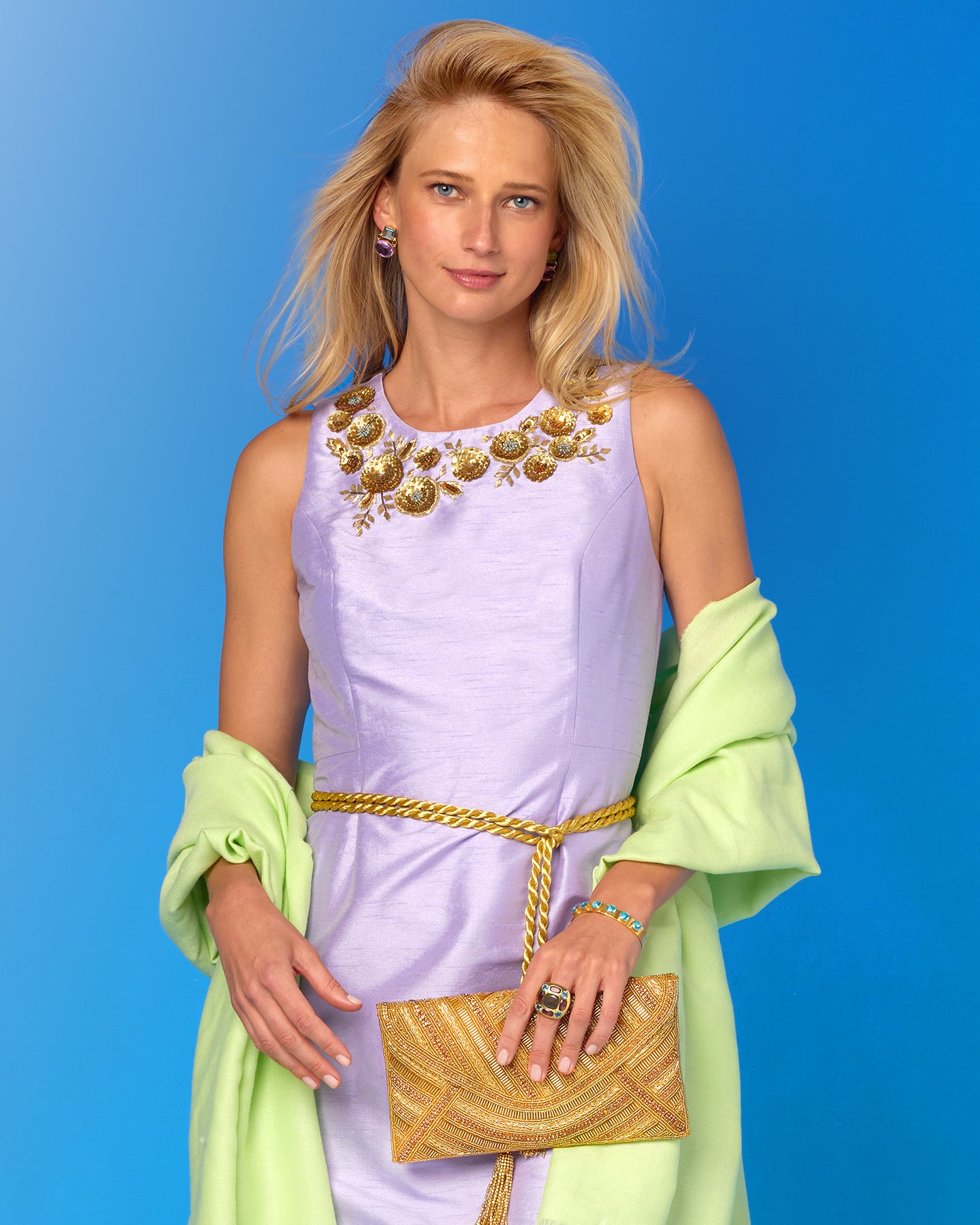 Woman in a light purple dress with gold accents and a green cloak holding a gold clutch against a blue background
