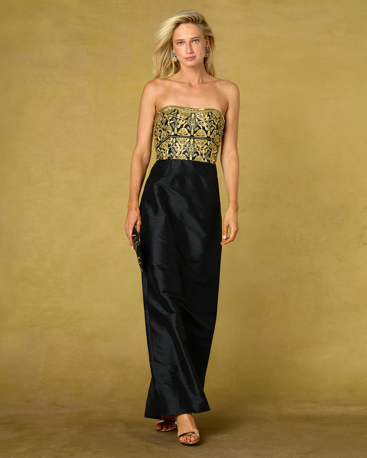 A woman models a strapless black gown with a wide, heavily embroidered gold and black patterned bodice, standing against a textured mustard-yellow studio backdrop.