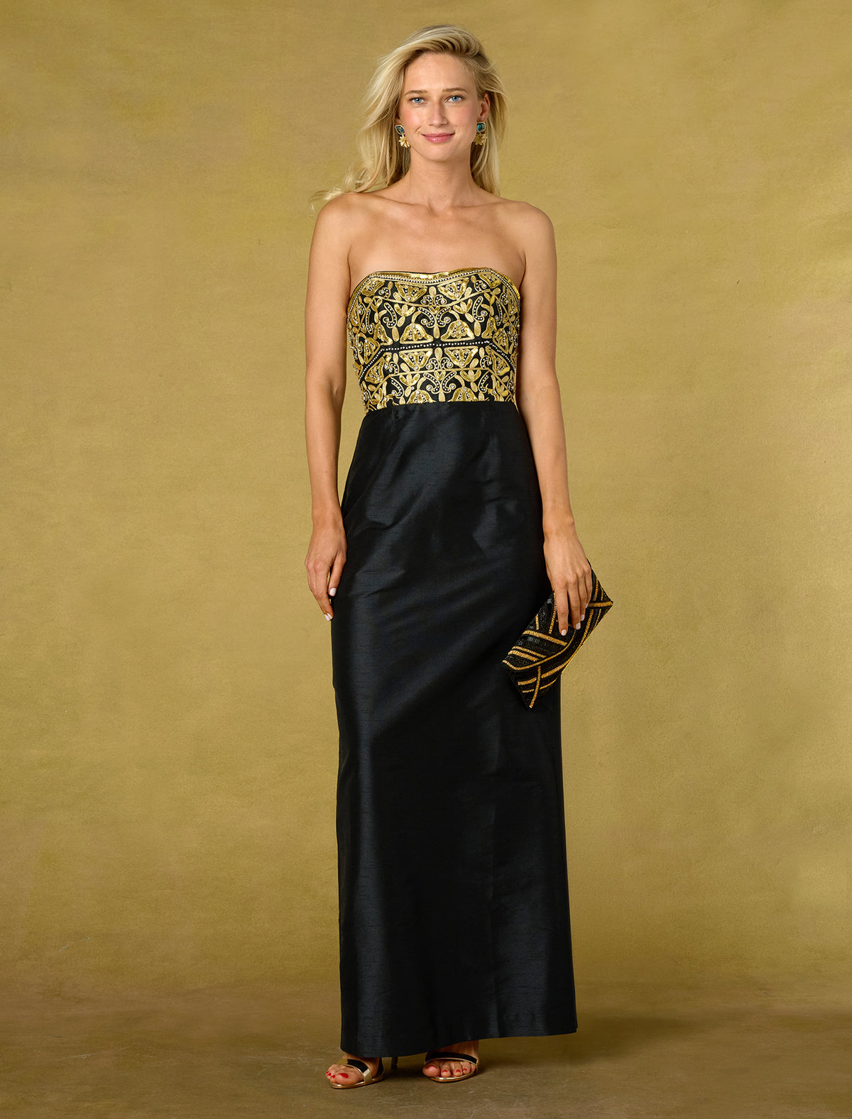 A woman models a strapless formal gown with a black satin skirt and a gold embroidered bodice, holding a matching black and gold clutch against a textured gold studio backdrop.