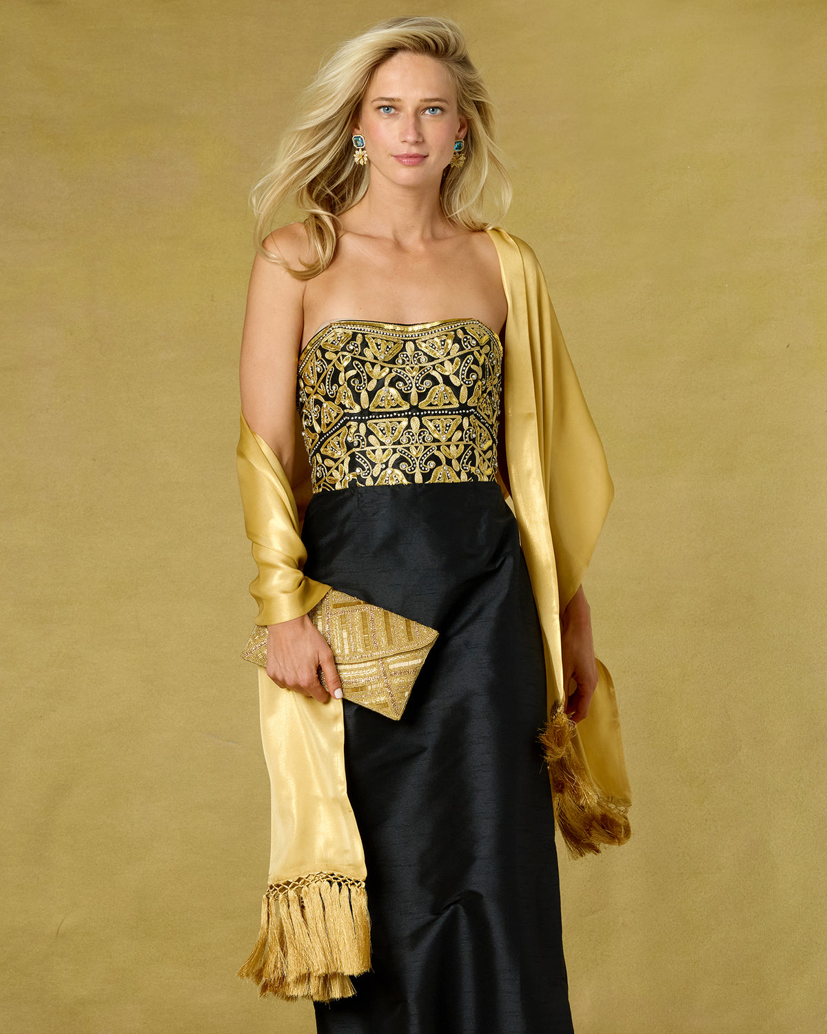 A blonde model wearing a strapless black gown with an ornate gold embroidered bodice, a matching gold draped shawl, and holding a gold clutch against a textured gold background.