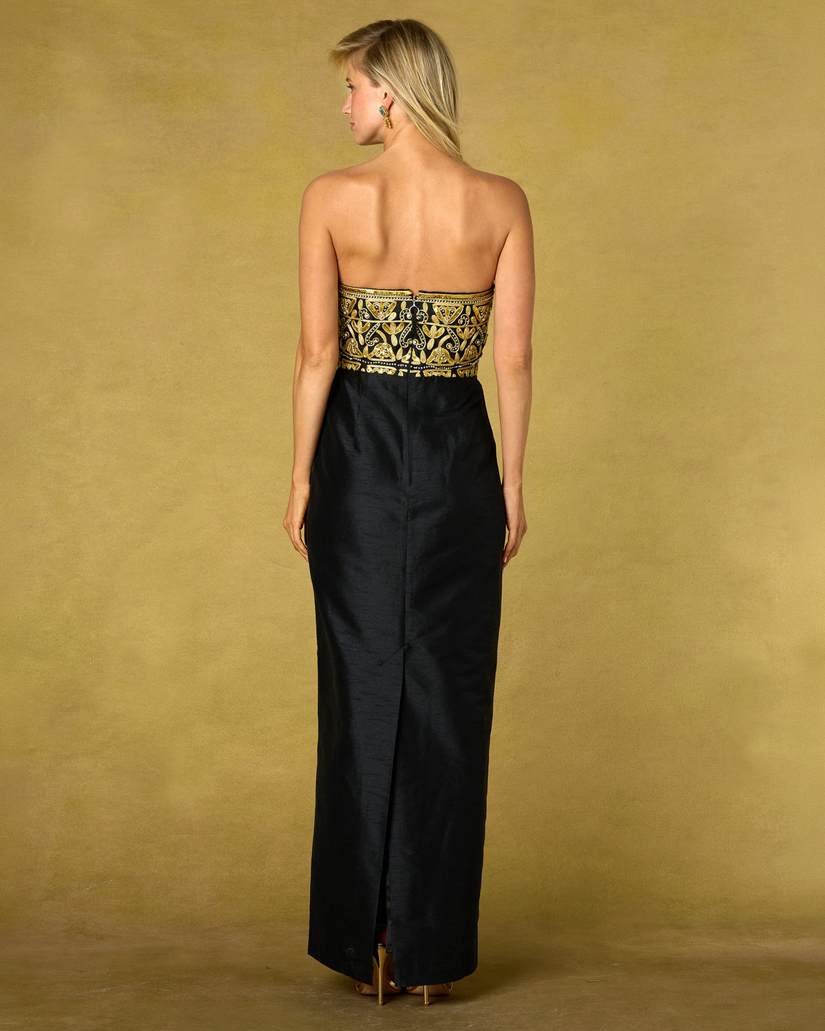 The back of a woman standing with her back to the camera wearing a strapless black gown with an ornate gold and black embroidered waistband, set against a gold backdrop.
