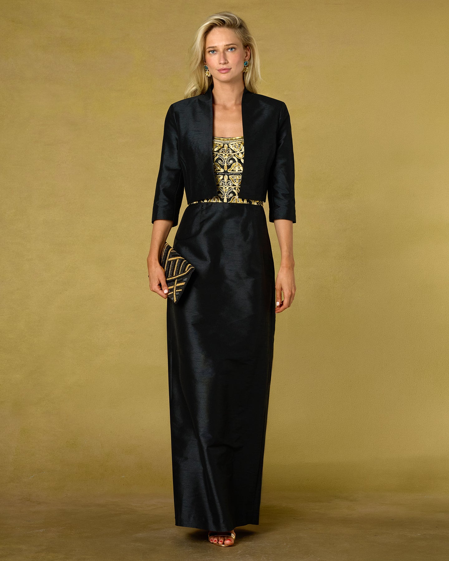 A woman modeling a black satin-like maxi dress with a gold embroidered bodice and a matching black blazer, standing against a warm olive-gold studio backdrop.
