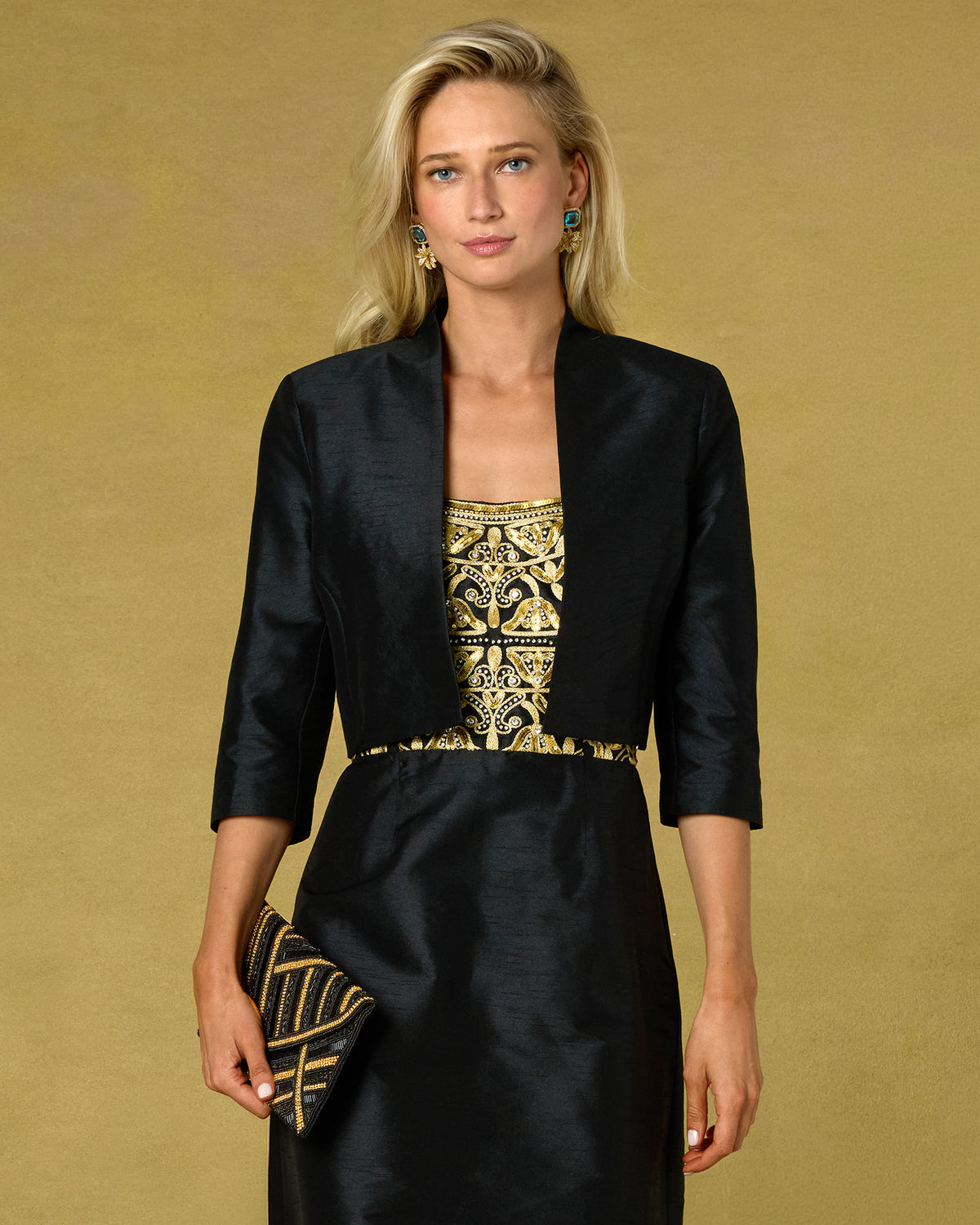 A blonde model wearing a black three-quarter sleeve jacket over a black dress with a gold embroidered bodice, holding a black and gold patterned clutch against a solid gold background.