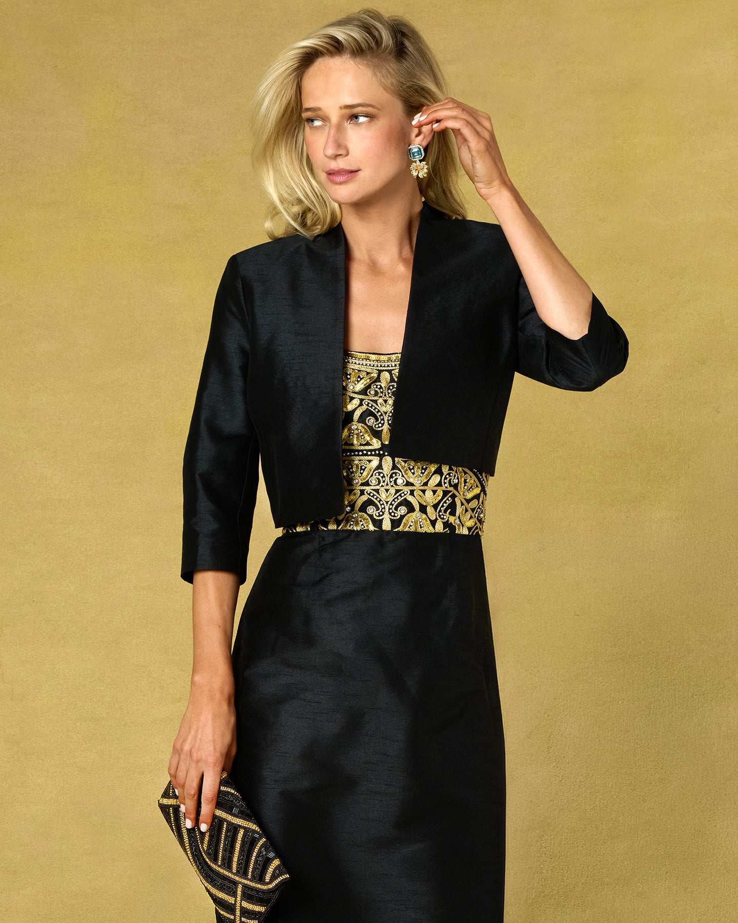 A blonde woman models a black three-piece matching set consisting of a cropped jacket, a patterned top, and a skirt, paired with a matching black and gold clutch against a textured gold background.