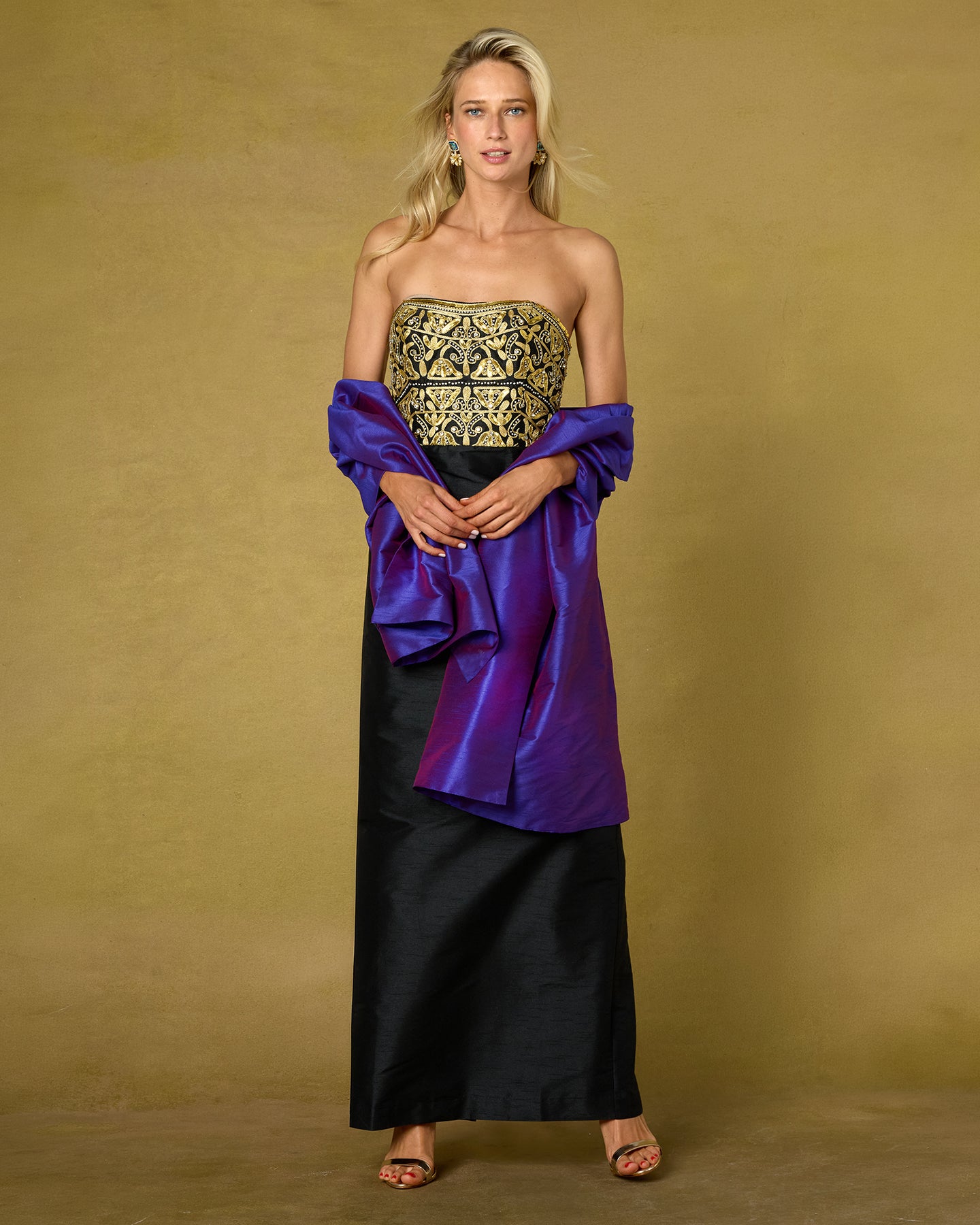 A blonde model wearing a strapless black gown with an ornate gold embroidered bodice and a draped purple satin cape, posed against a textured gold background.