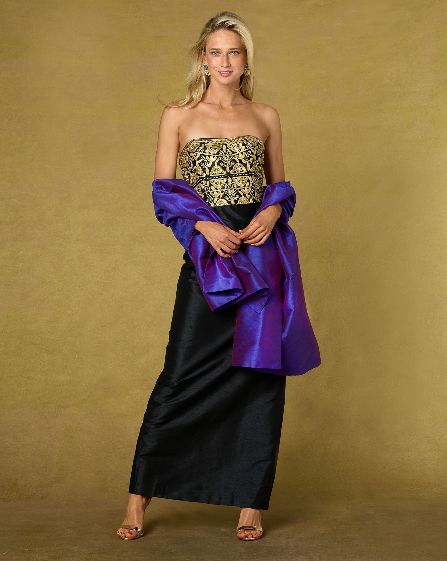 A woman models a strapless black taffeta dress with a gold-embroidered bodice and a draped purple taffeta sash, set against a solid gold studio backdrop.