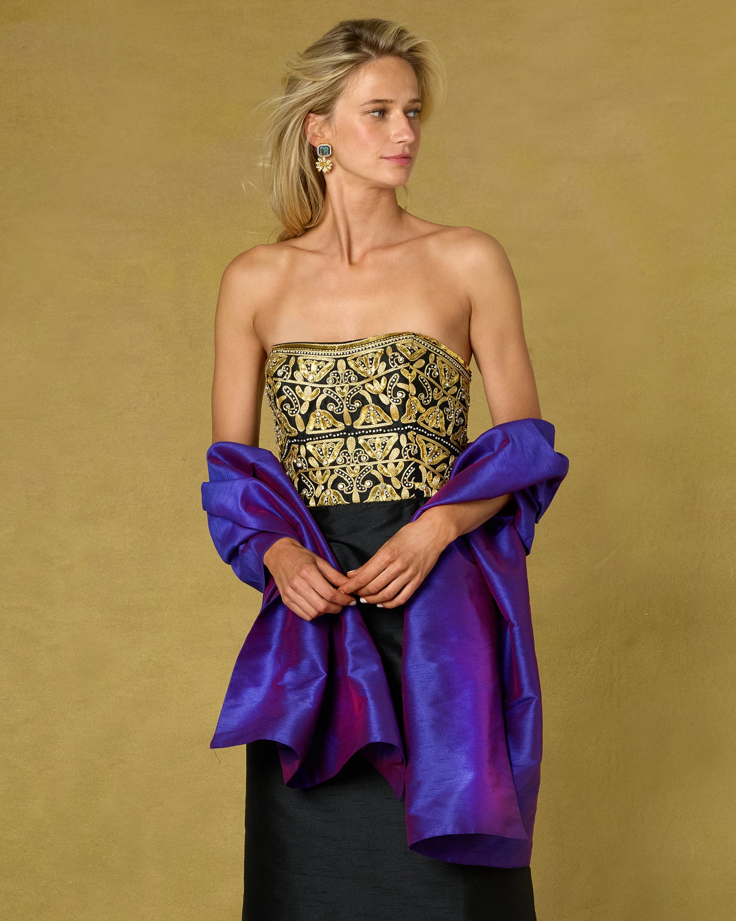 A model wearing a strapless black and gold embroidered tube top with voluminous purple draped fabric sleeves, paired with a black skirt, against a textured gold background.