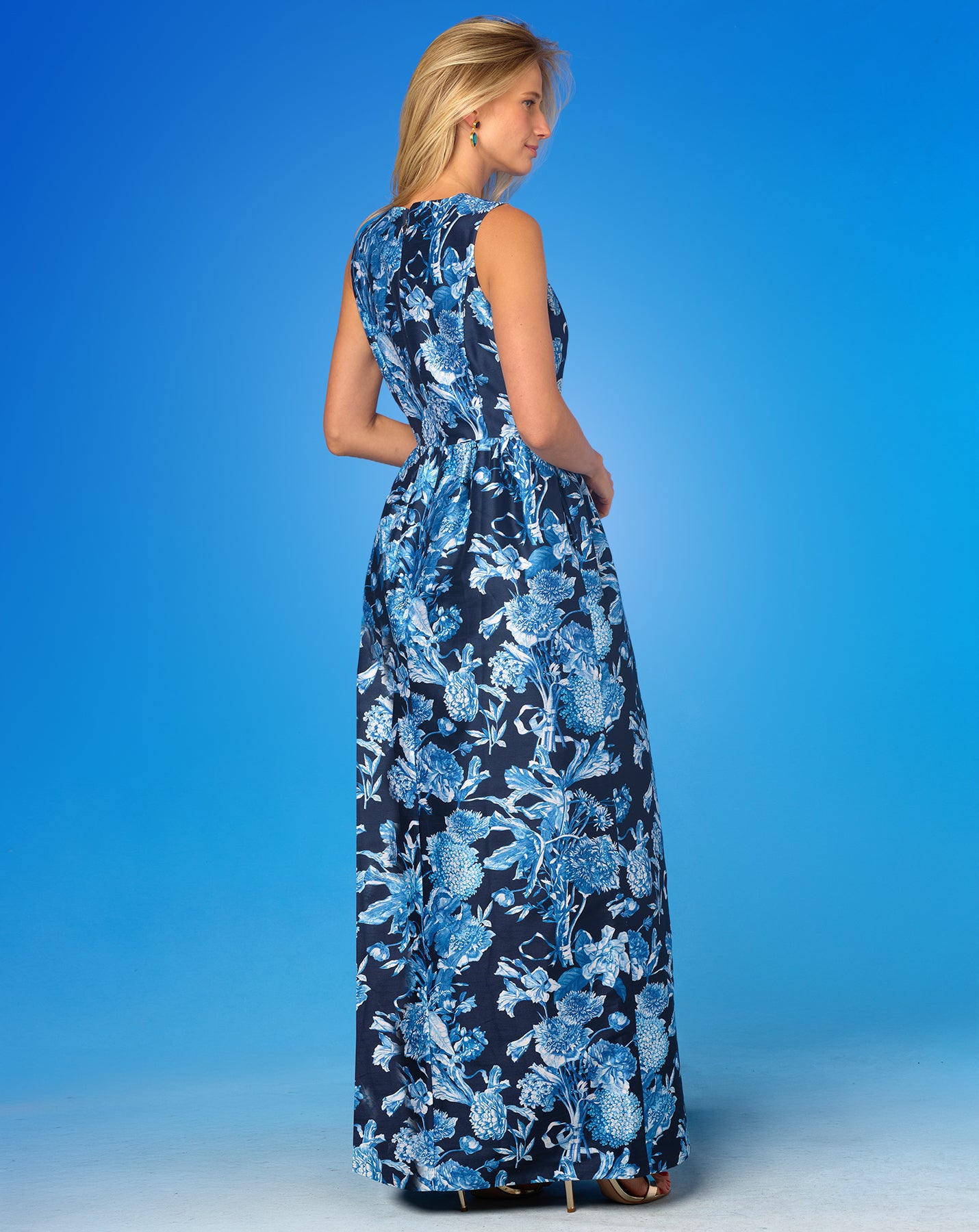 Back view of a woman wearing the Elodie Long Dress in Camille Navy and White Floral Toile against a blue background