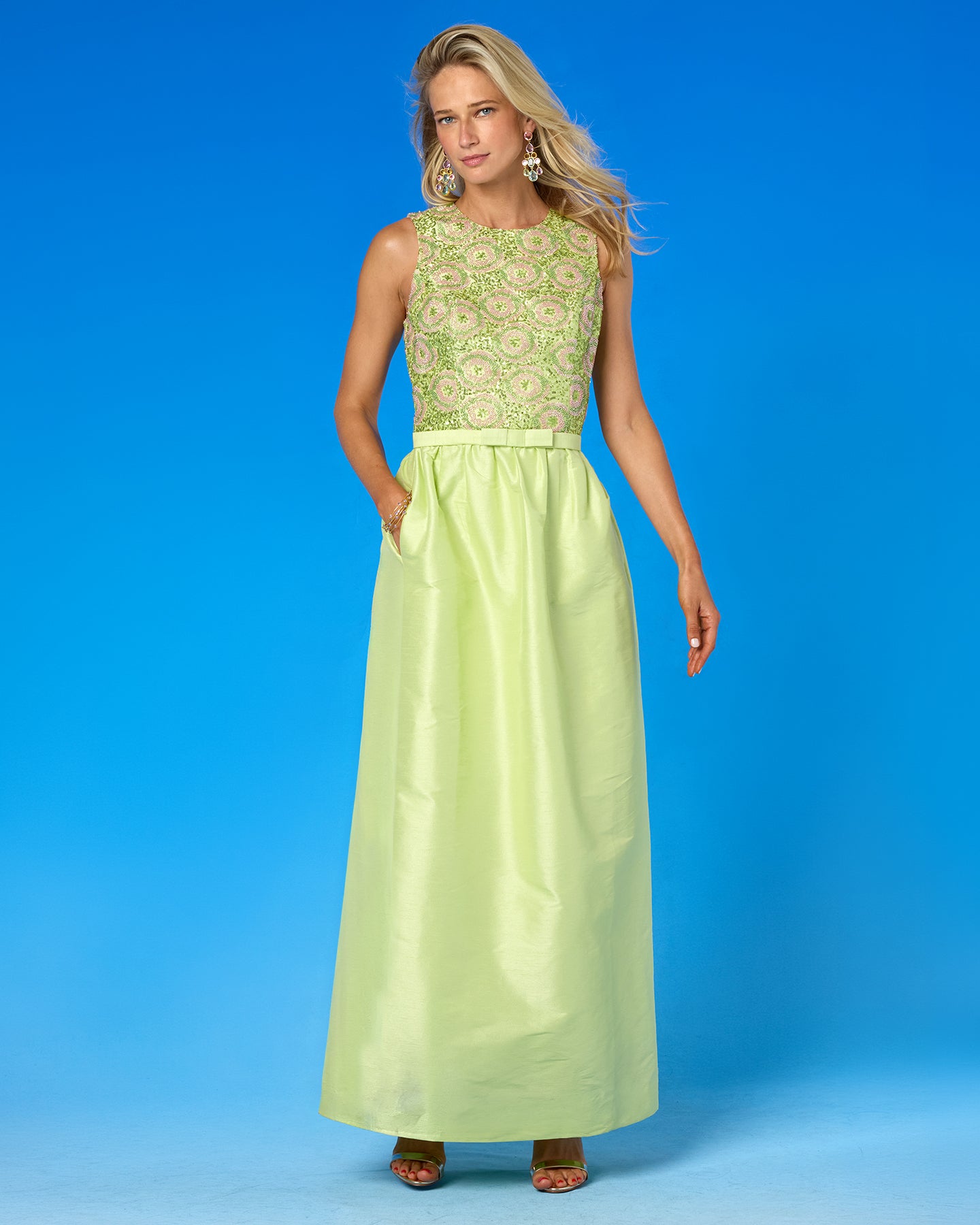 A woman modeling a sleeveless floor-length dress with a light green and white floral lace bodice and a solid pale yellow-green satin skirt, set against a solid blue studio background.