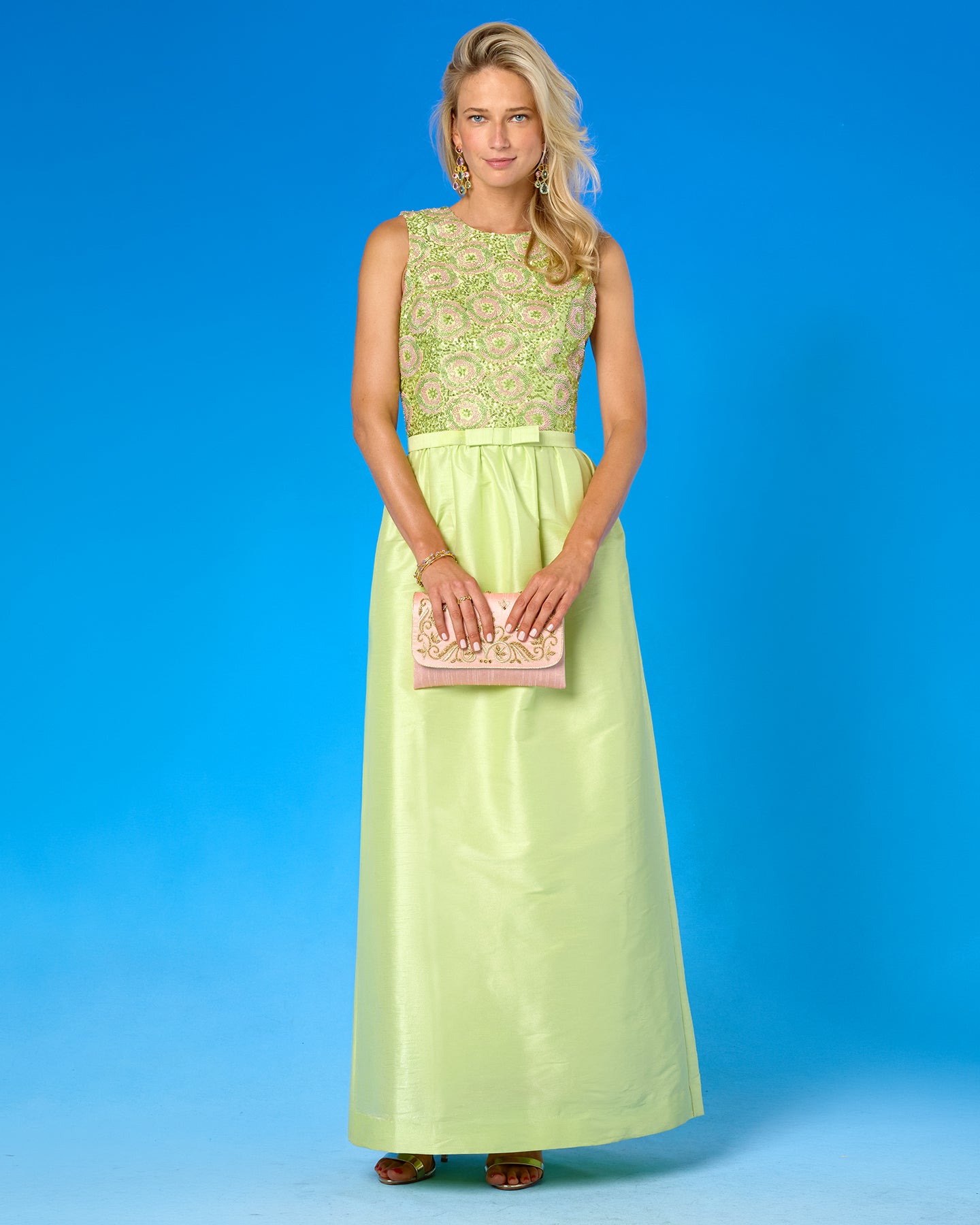 A woman modeling a sleeveless maxi dress with a light green satin skirt and a beige and green embroidered bodice, holding a matching clutch bag against a solid blue studio background.