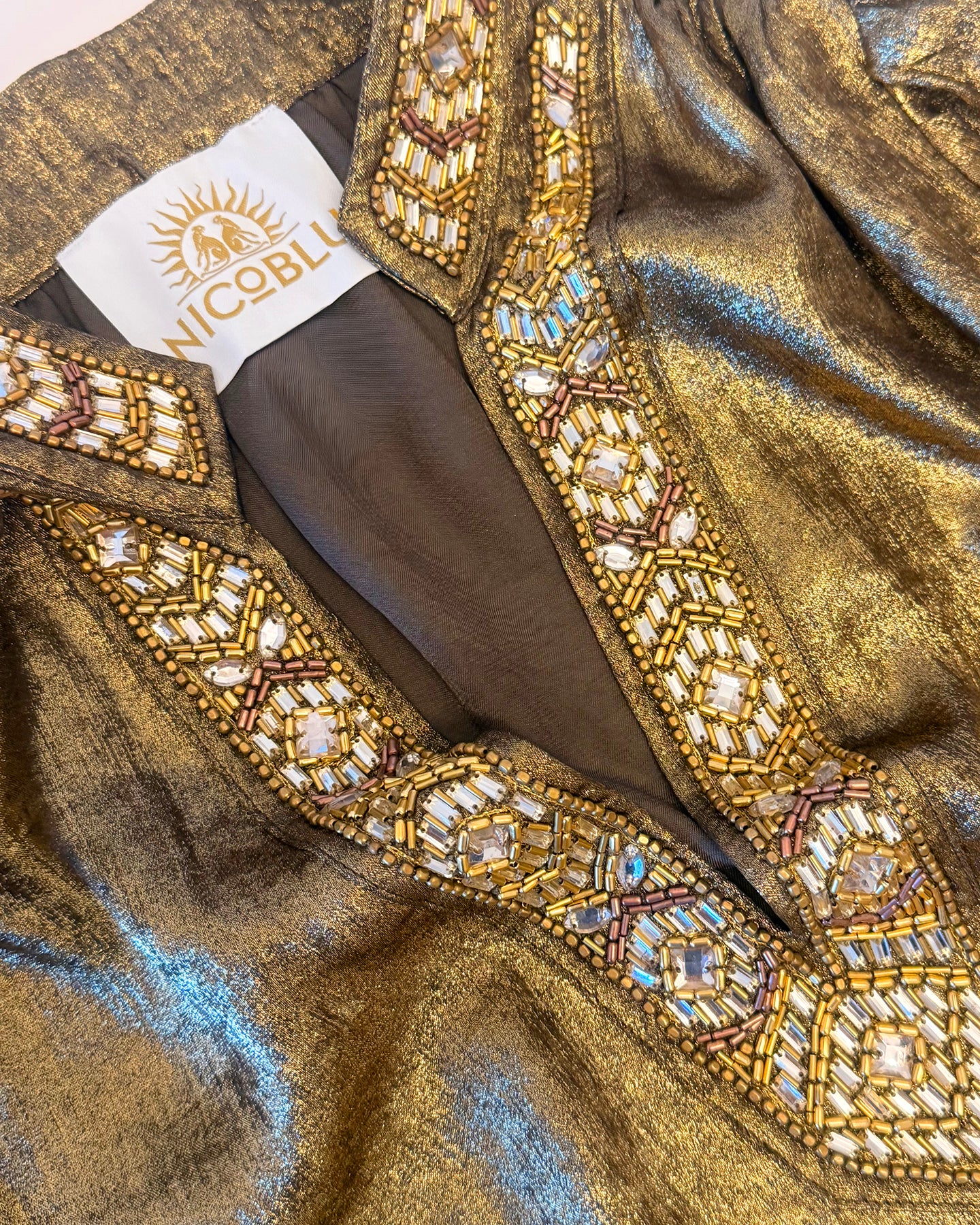 Close-up of a metallic gold fabric garment with ornate multicolor beaded embroidery around a deep V-neckline. A white brand label reading NICOBLOO is visible inside the neckline.