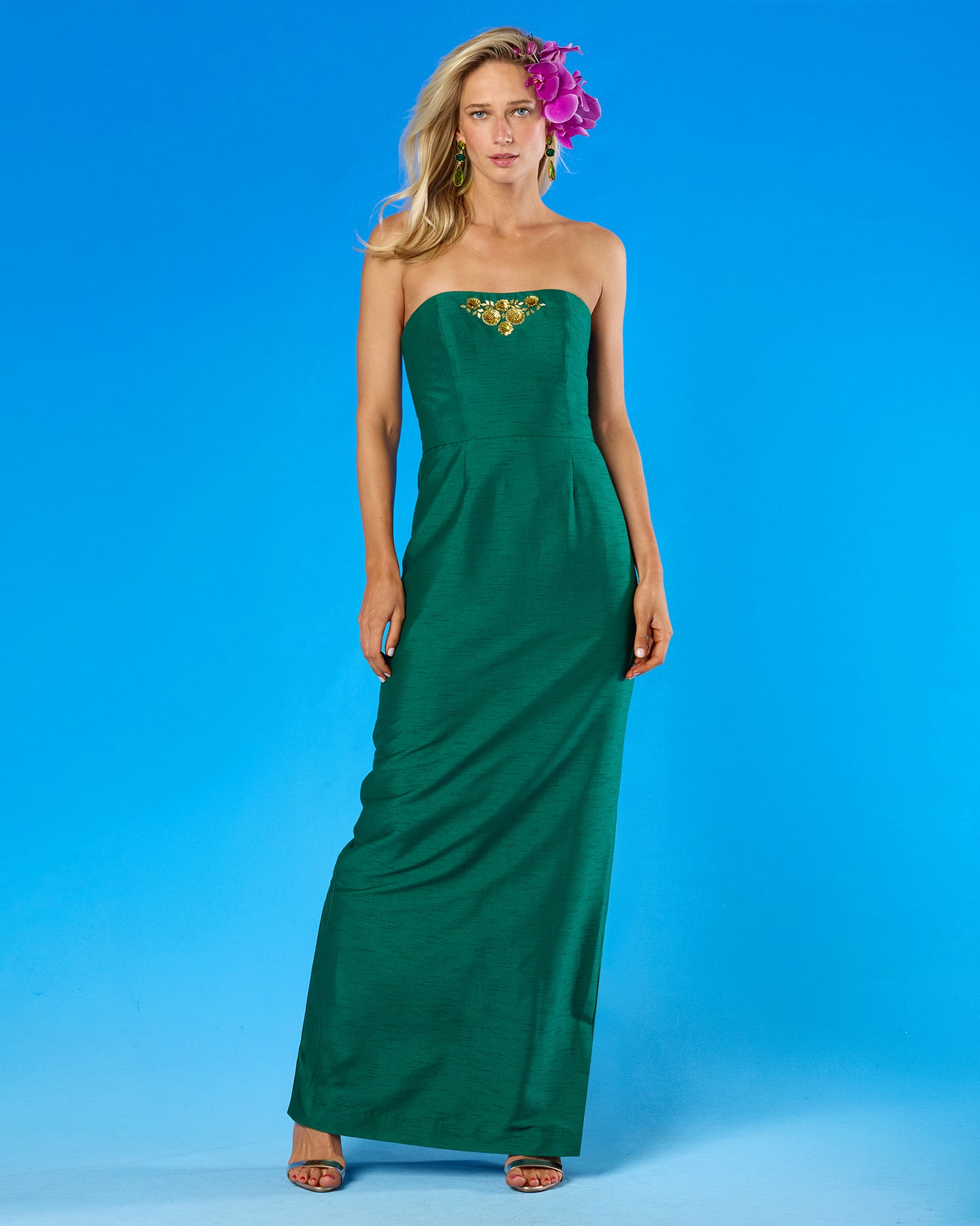 A woman modeling a strapless emerald green maxi dress with a gold embroidered chest detail, standing against a solid blue studio background.