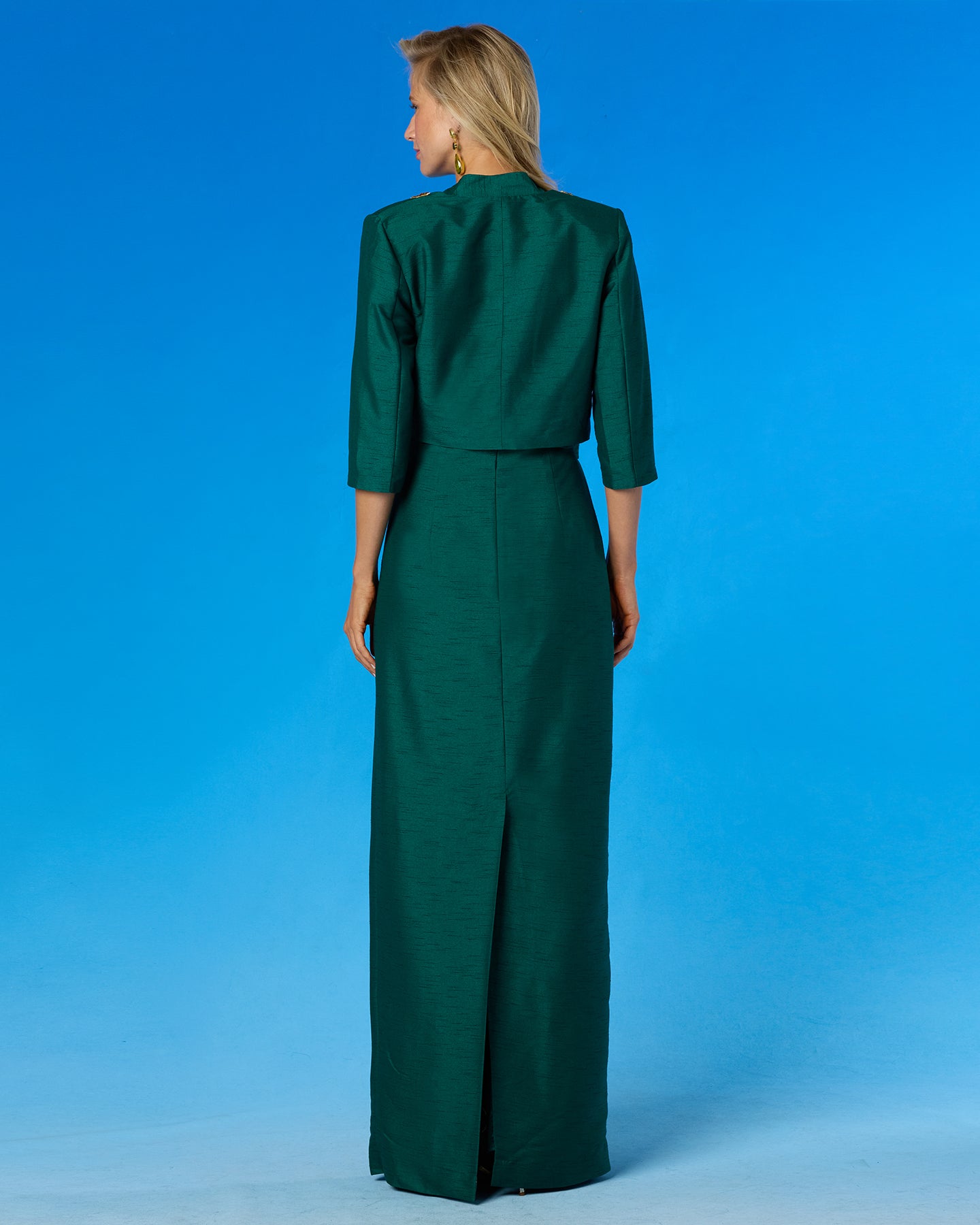 A woman viewed from the back wearing a dark green two-piece outfit consisting of a cropped jacket with three-quarter sleeves and a matching long skirt, set against a solid blue studio background.