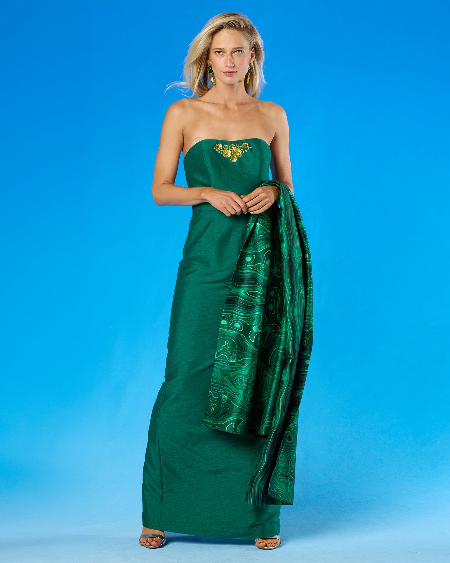 A woman modeling a strapless emerald green saree with a gold embroidered bodice and a matching patterned green and gold border drape, set against a solid blue studio background.