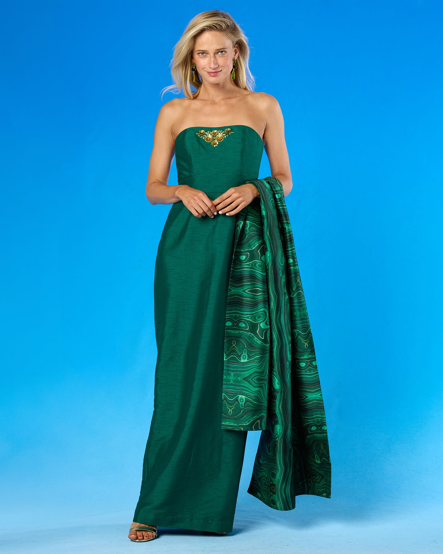 A woman modeling a strapless dark green gown with a gold embellished neckline and a matching patterned green dupatta, set against a solid blue studio background.